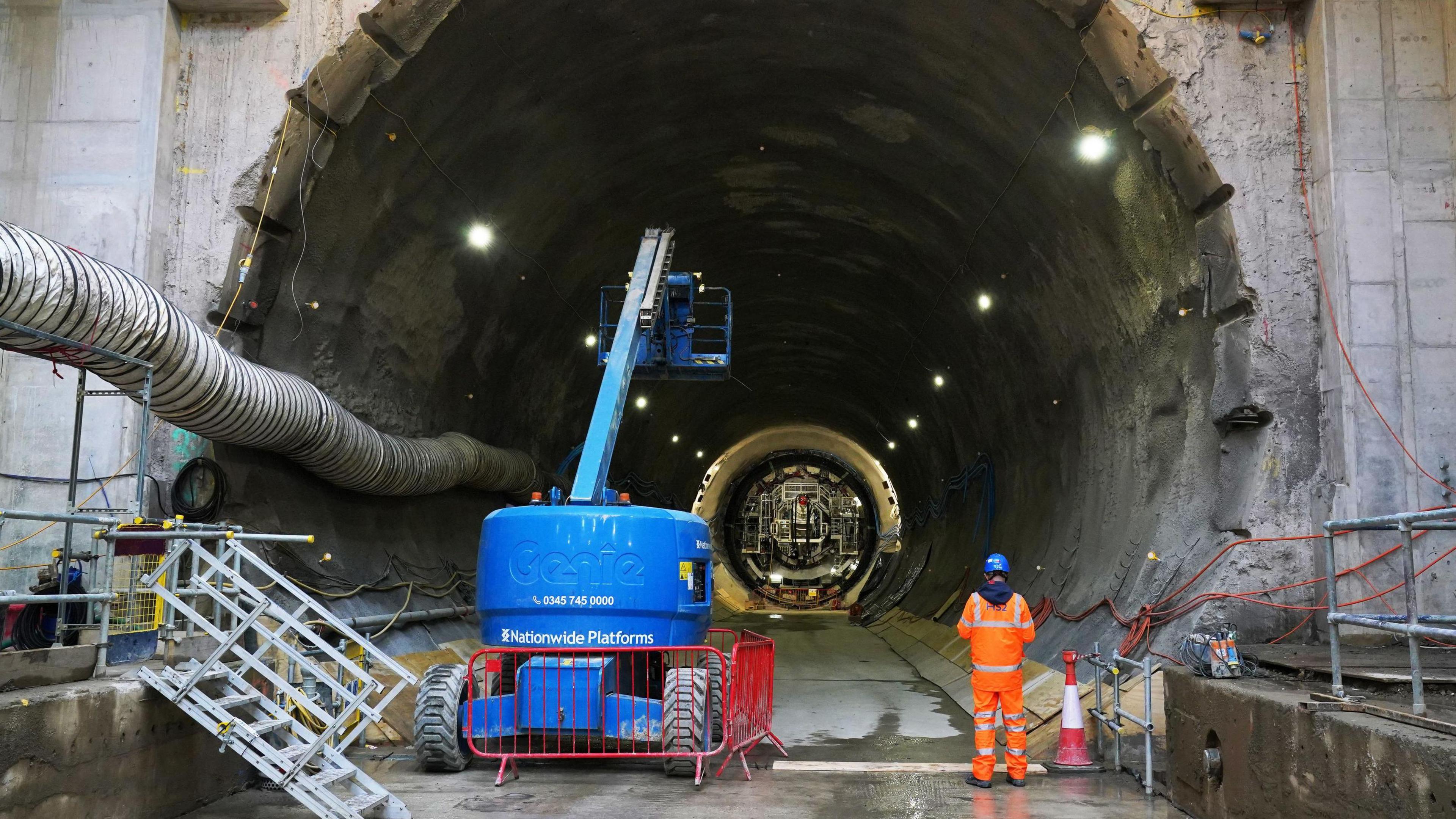 A large tunnel with one of the boring machines in the background, with a cherry picker and a construction worker in orange high vis and a blue hardhat in the foreground. 