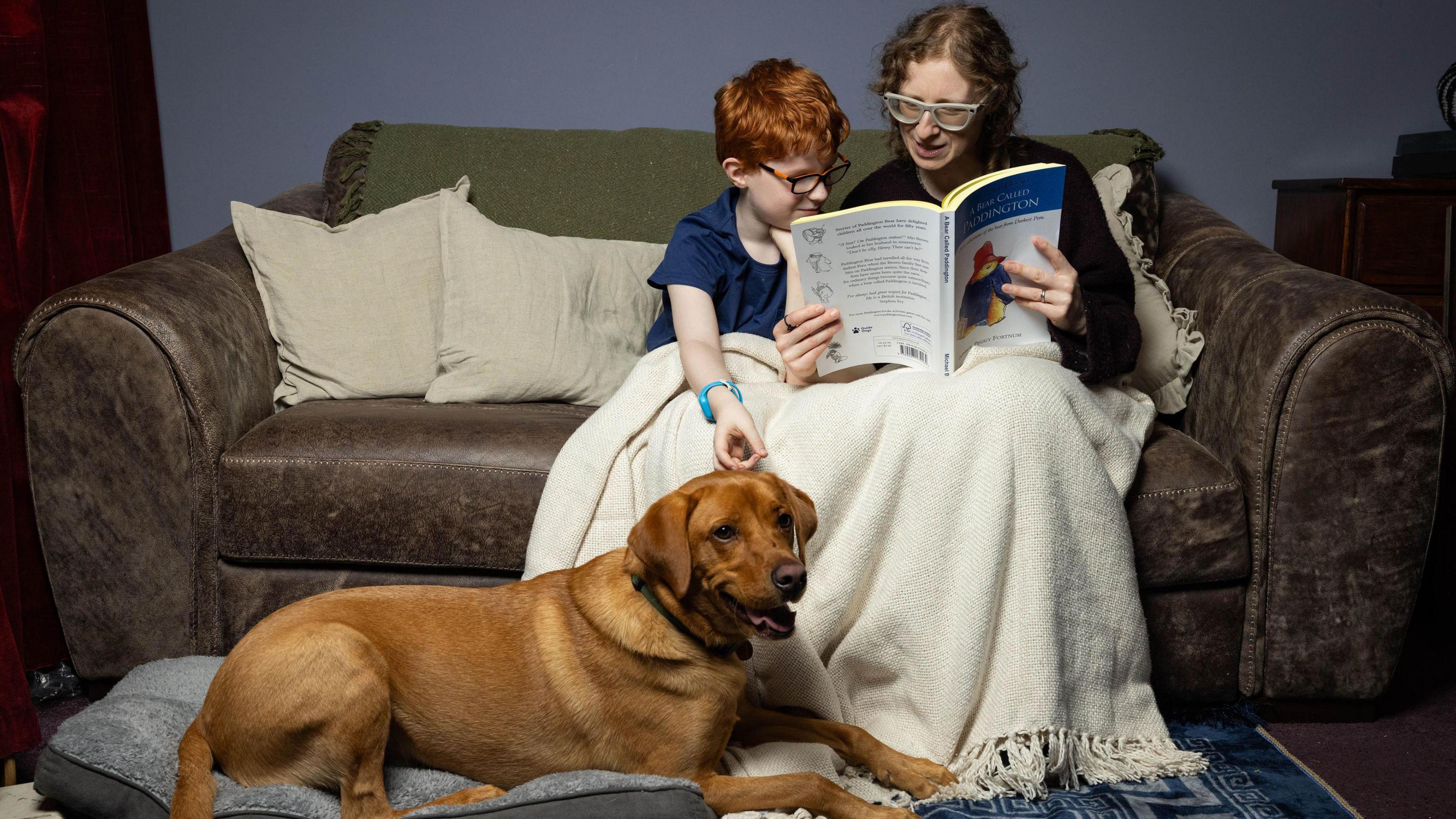 A boy with red hair and a woman sit on a brown sofa with a labrador at their feet. Both mum and son are wearing glasses, and the woman is holding a book which they are both looking at.  They are sitting under a cream throw.