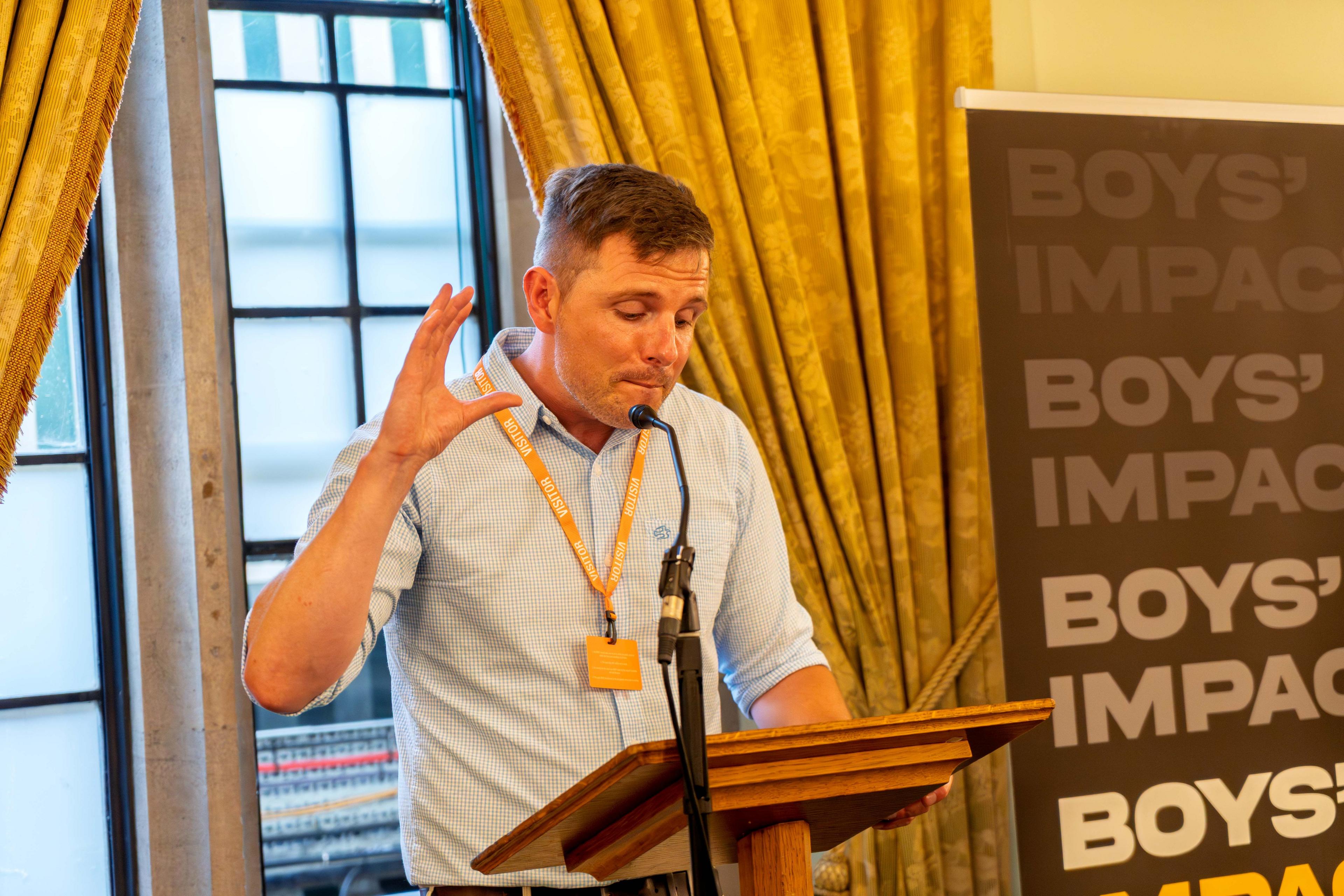 Dr Alex Blower is lasting  astatine  a woody  lectern speaking into a microphone. He is wearing a bluish  checked garment  with his sleeves rolled up   and is looking down   astatine  notes portion    helium  talks into the microphone. Behind him is simply a interaction   with the Boys' Impact logo. 