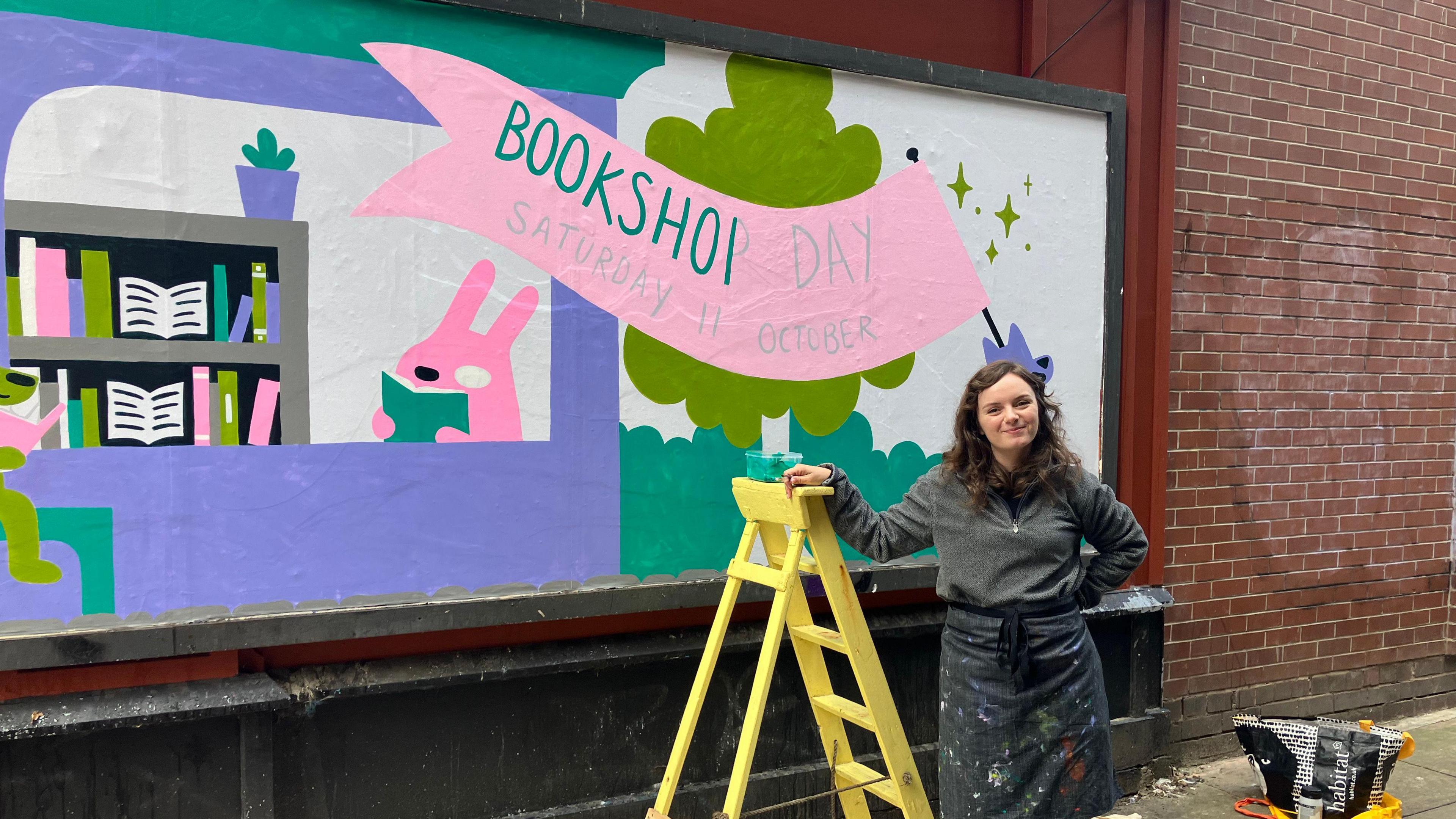 A person stands beside a yellow ladder in front of a vibrant mural promoting 'Bookshop Day' on Saturday 11 October. The mural features colourful illustrations of bookshelves, books, and cartoon animals, with a prominent pink banner displaying the event details. Artist Bethan Woollvin is dressed in a dark apron and holding paint supplies. Painting tools and materials are scattered on the ground nearby.
