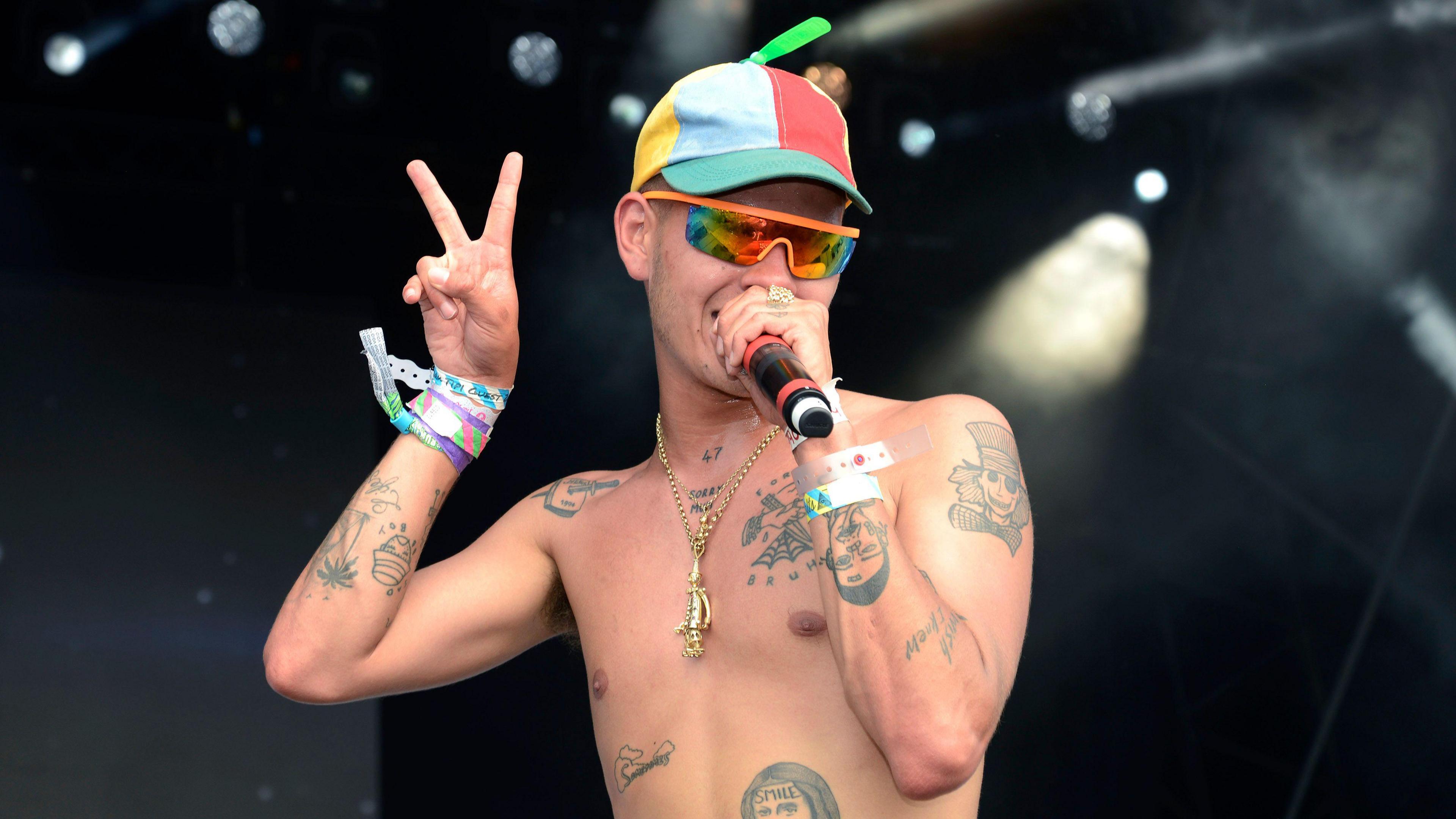 Slowthai performing at a festival. He is wearing a multicoloured hat and sunglasses and is singing into a microphone. He is shirtless, showing several tattoos on his torso and arms. He is making a peace sign with his right hand.