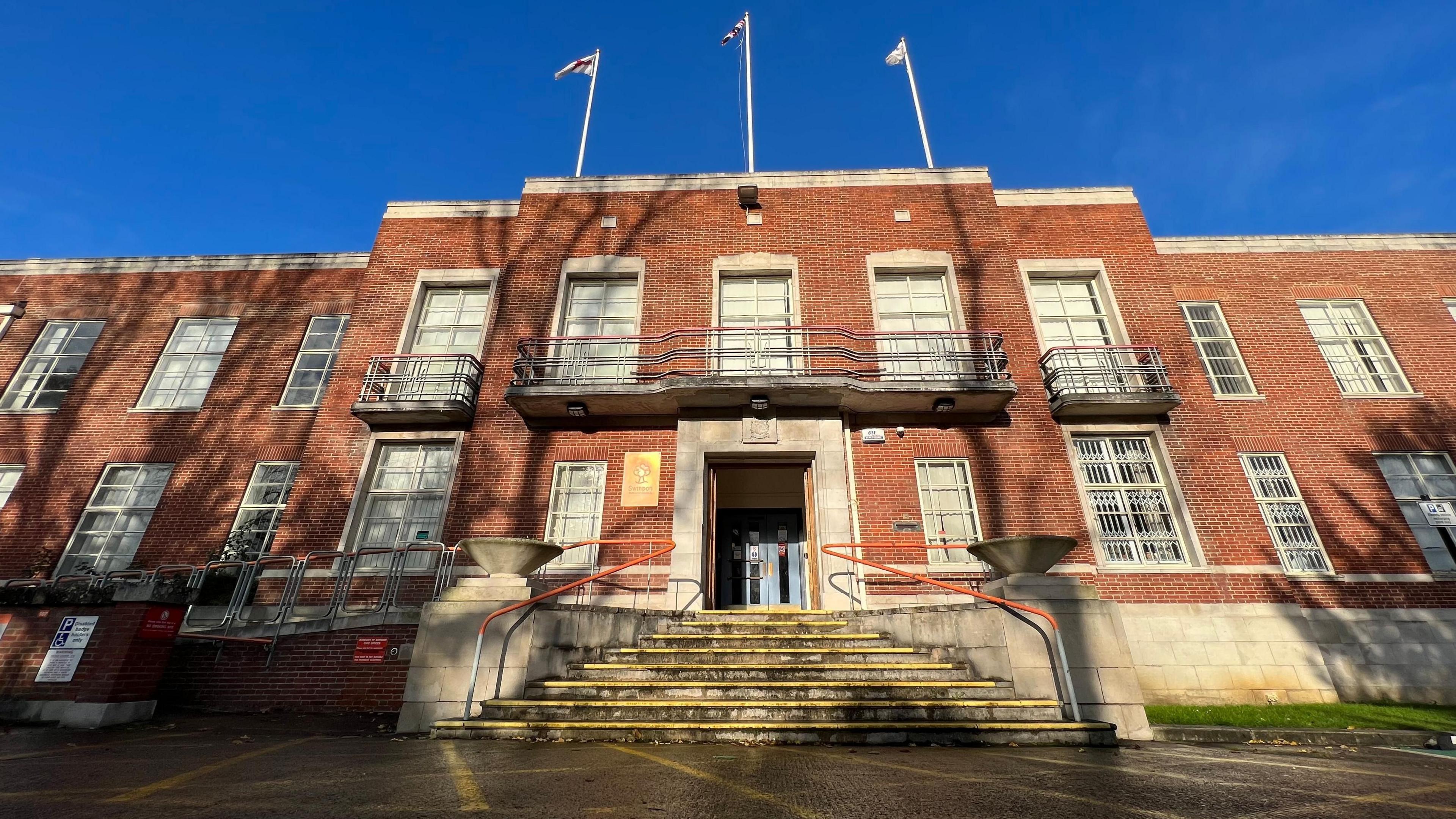 The outside entrance to Swindon Borough Council's headquarters on a sunny day with a blue sky and three flags on its roof