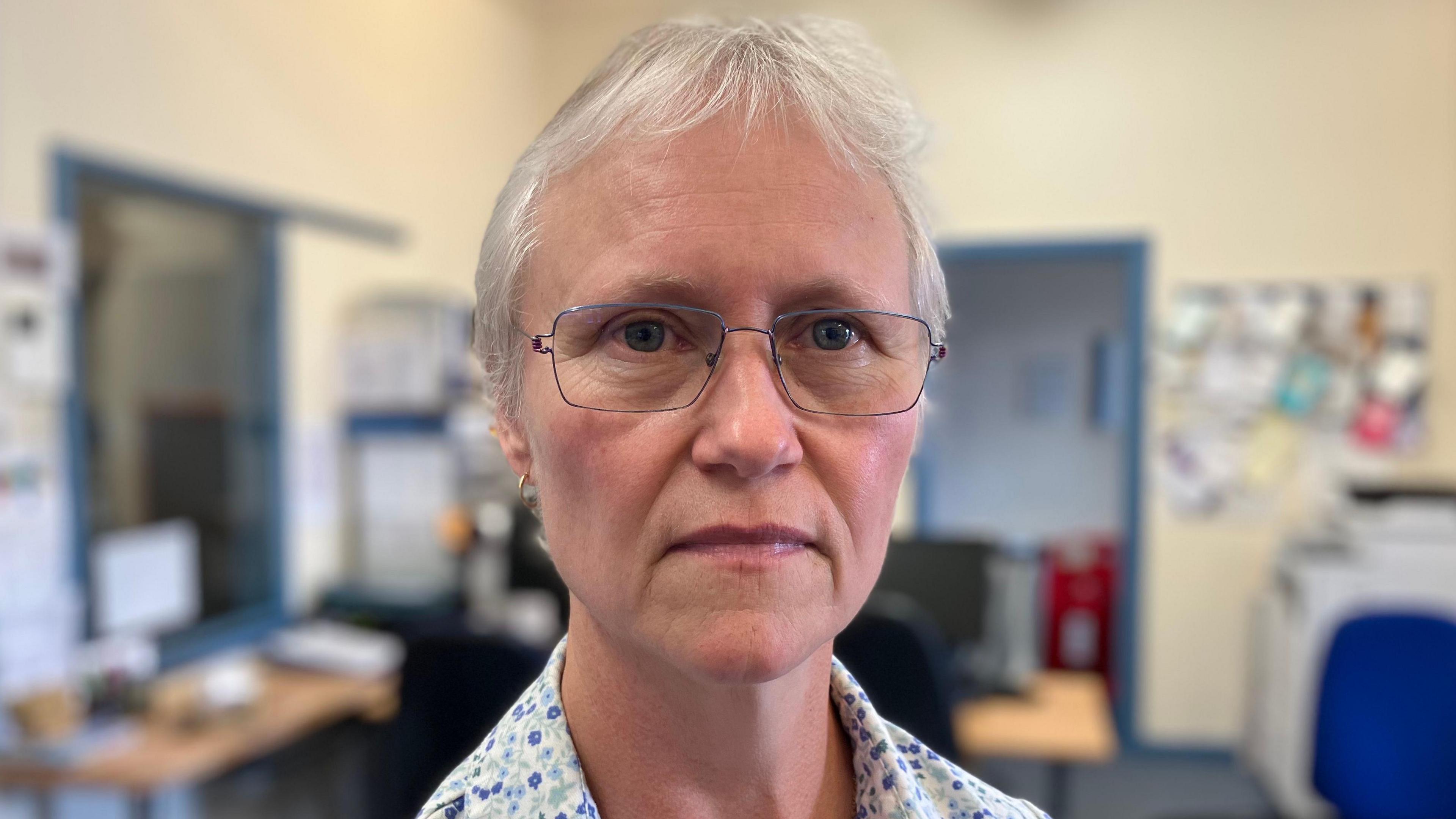 Pat Coomber-Wood is looking at the camera in an office with a straight face while wearing a top with a flower design on it.