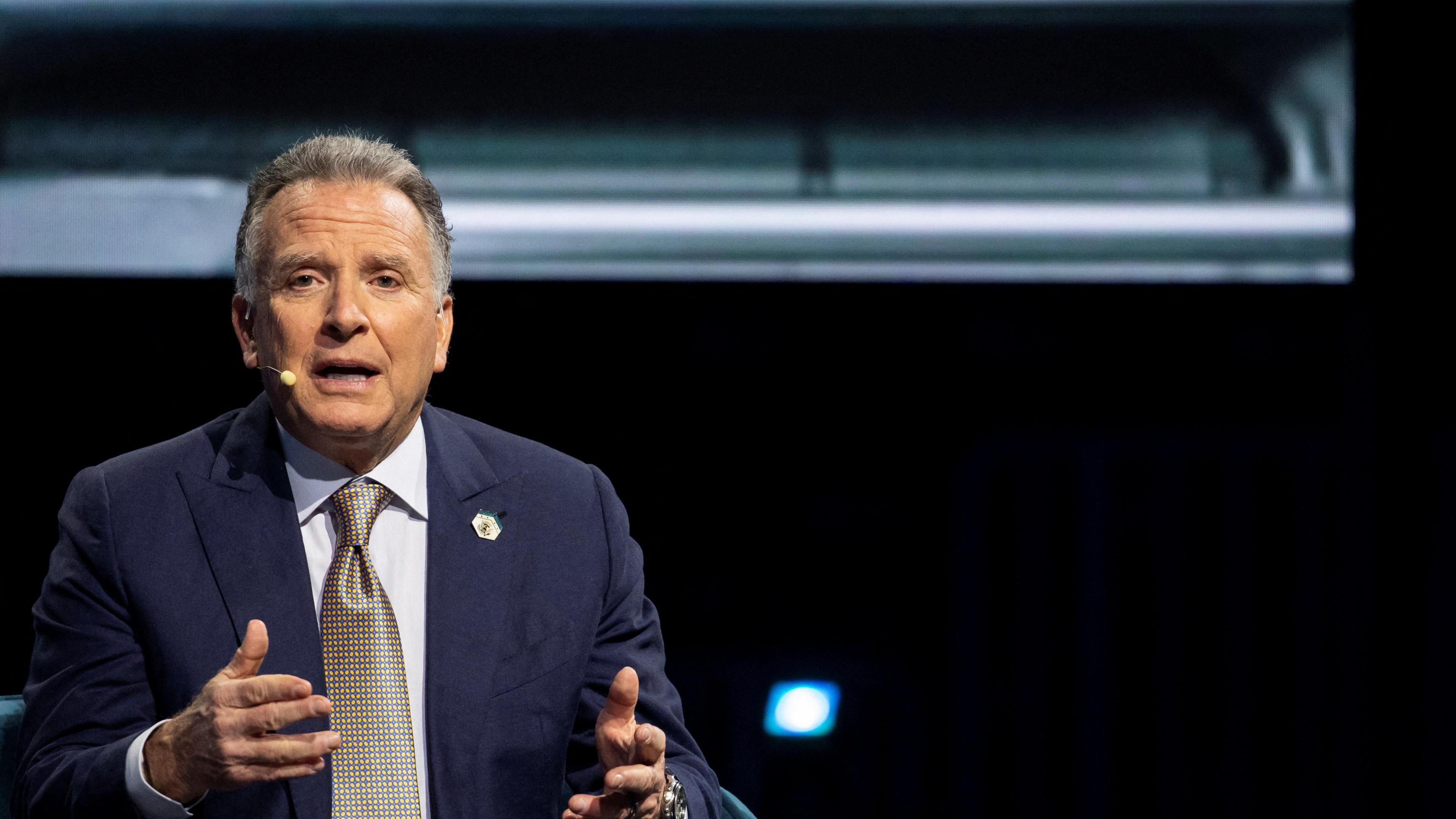 A middle-aged man in a blue jacket, white shirt and gold tie gesticulates in a room.  