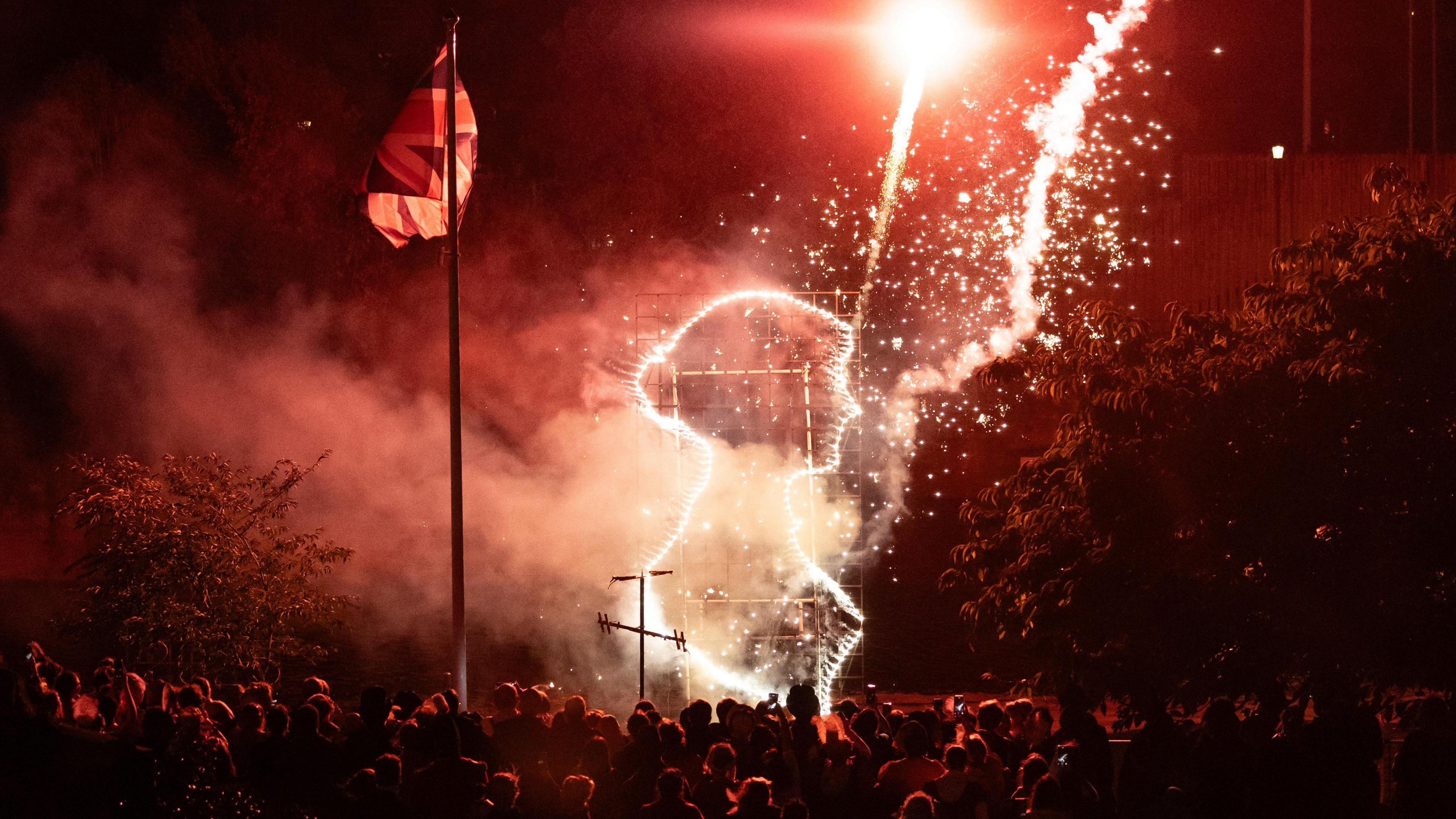 A shot taken at night time. There is a large crowd of people all facing a light display depicting the silhouette of Jane Austen. A union jack flag can be seen next to it and there are red fireworks shooting up to the sky.