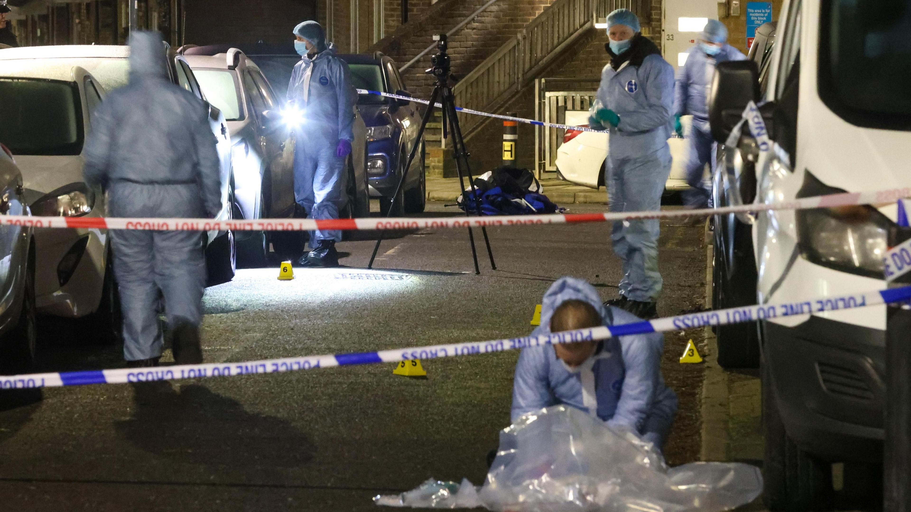 Forensic investigators in protective suits examine a cordoned-off crime scene on a residential street at night.