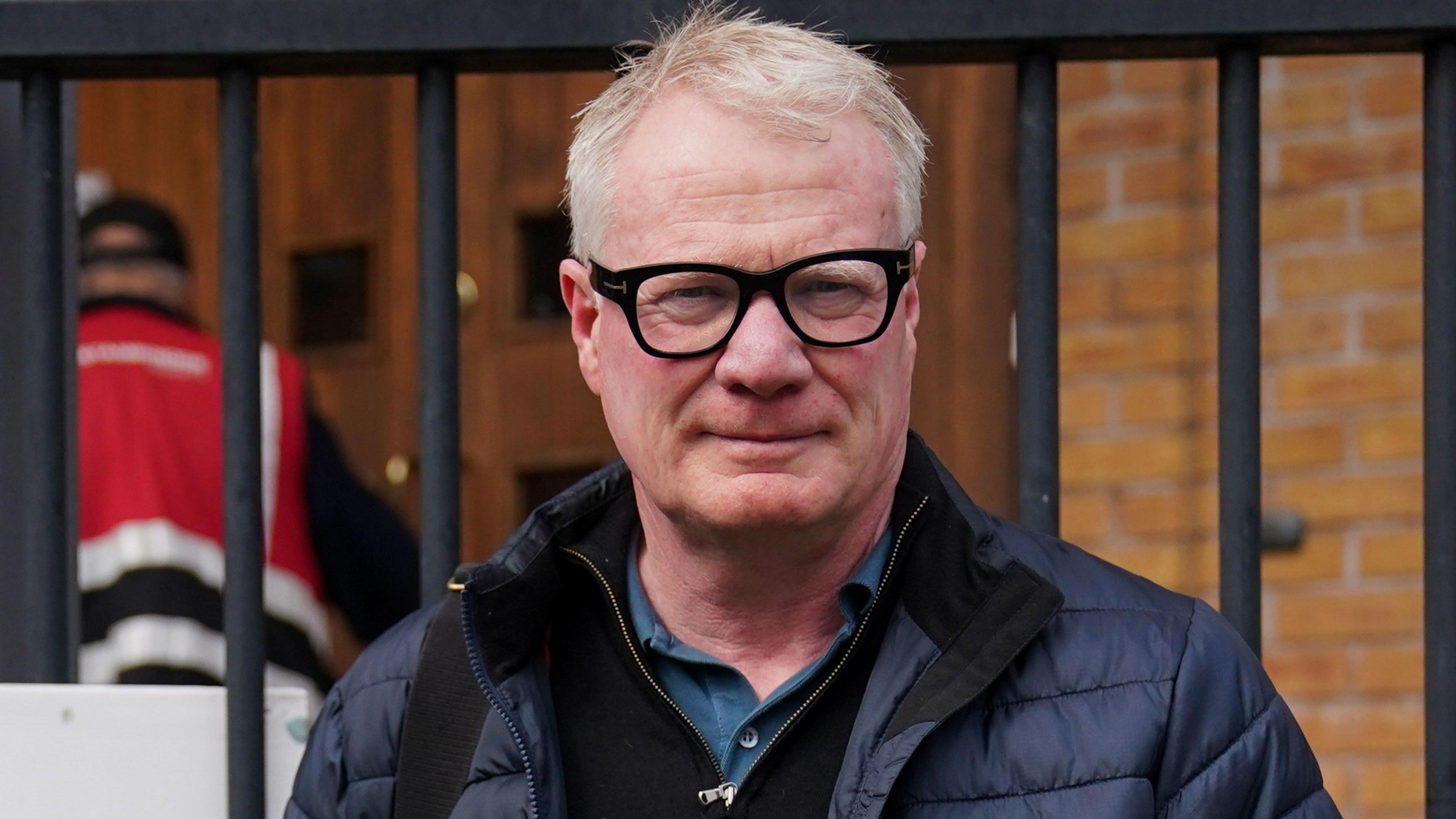 Richard Parker, wearing black glasses and a dark jacket, is stood in front of a black gate. He has short blonde, white hair.