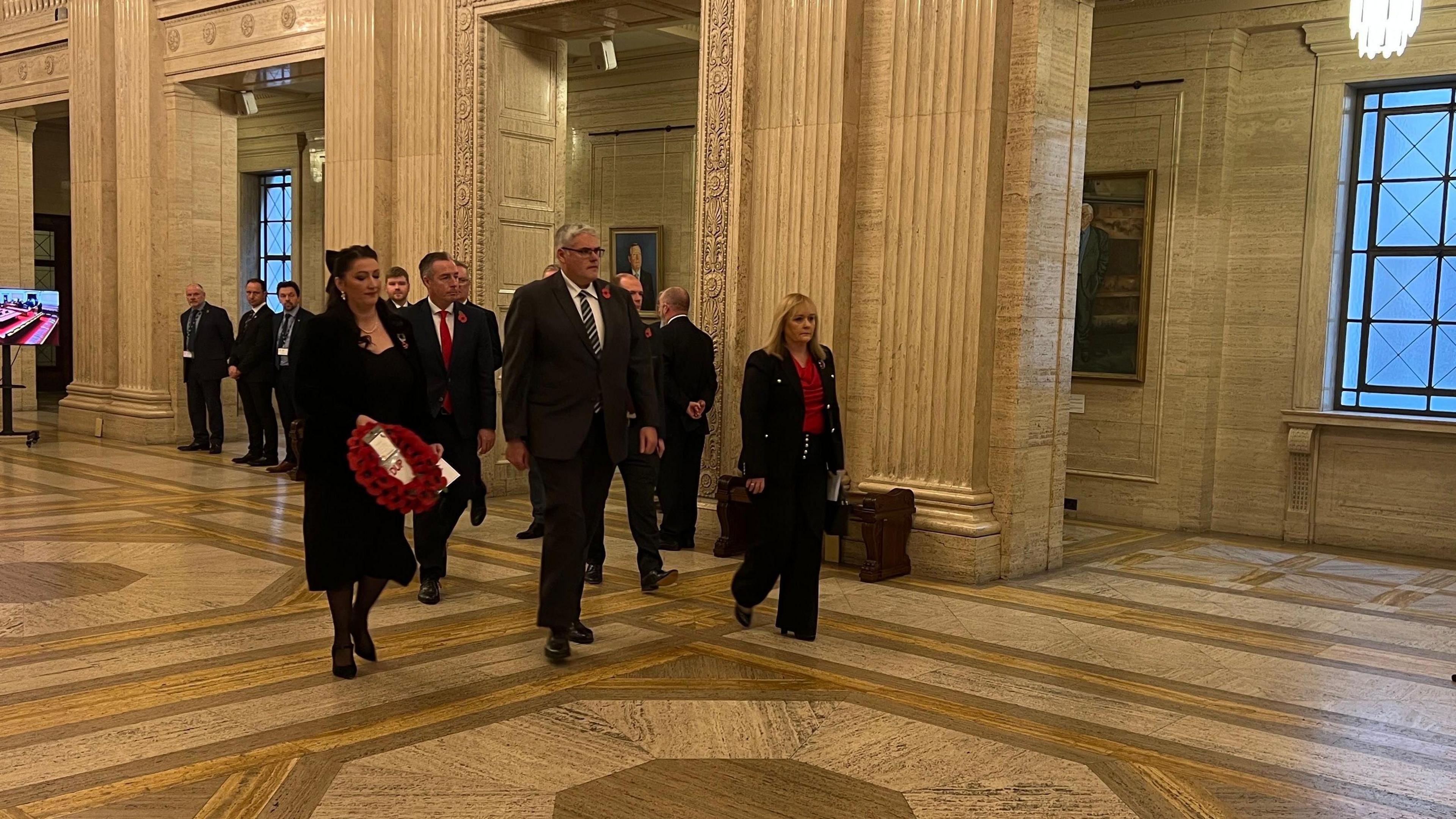 A man in formal attire with grey hair, glasses and a poppy walks through an official looking building. A woman to his right with black, also wearing formal attire and a poppy is holding a wreath. A blonde haired woman, also in formal attire and wearing a poppy is walking to his right. On lookers in formal attire can be seen in the background.   