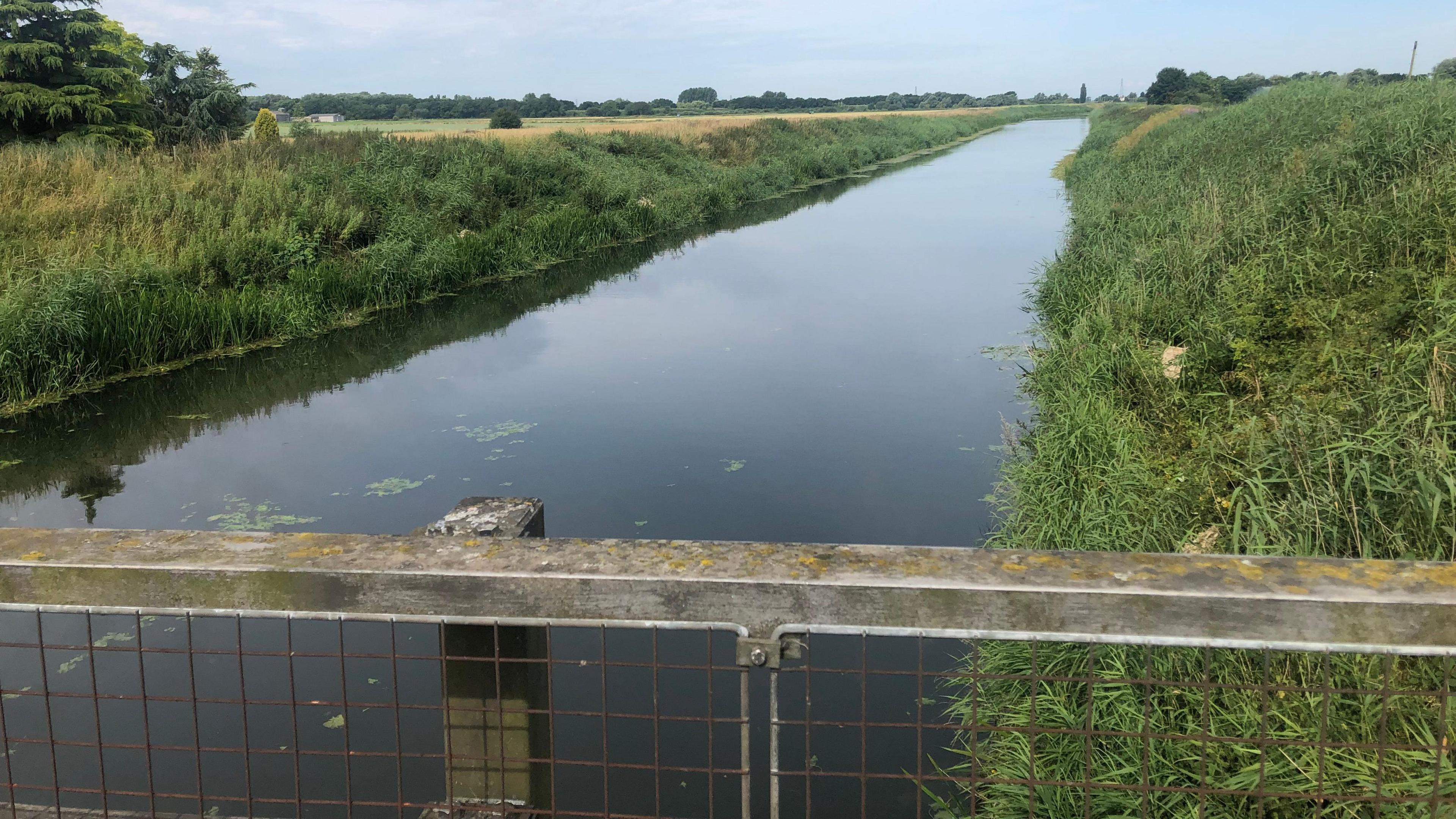 The river from the top of Chain Bridge, with grassy banks either side. The flat countryside landscape is in the distance.