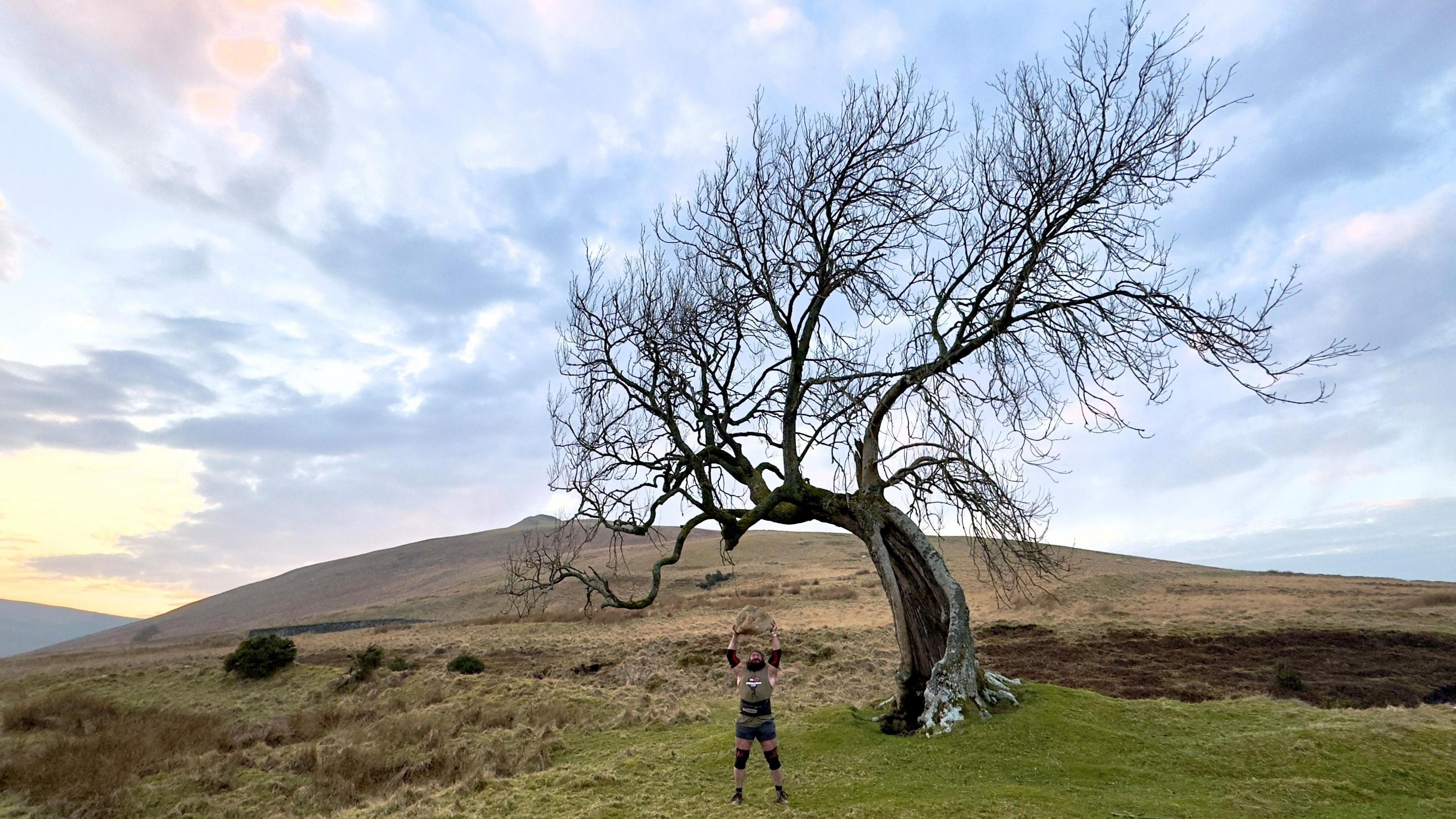 A bare tree in the middle of the image. A man is standing in the centre on a grassy, green floor. He is dressed in dark colours and is lifting a large stone above his head. The sky is bright but there are a number of light and dark coloured clouds overhead. There is a hill in the background.