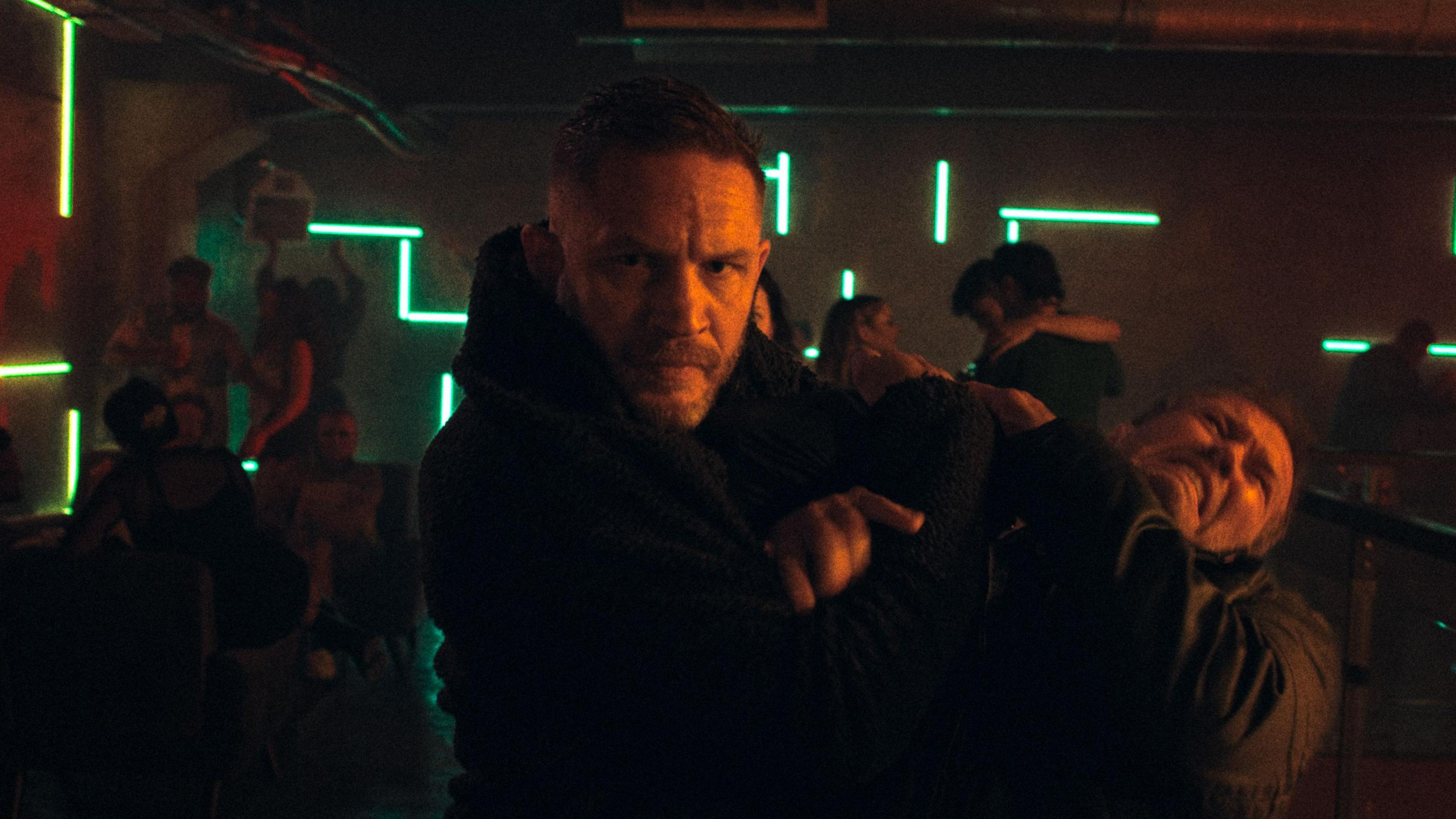 Tom hardy, who has another man in a chokehold. Both men are wearing all black and in the background is a nightclub with people dancing.