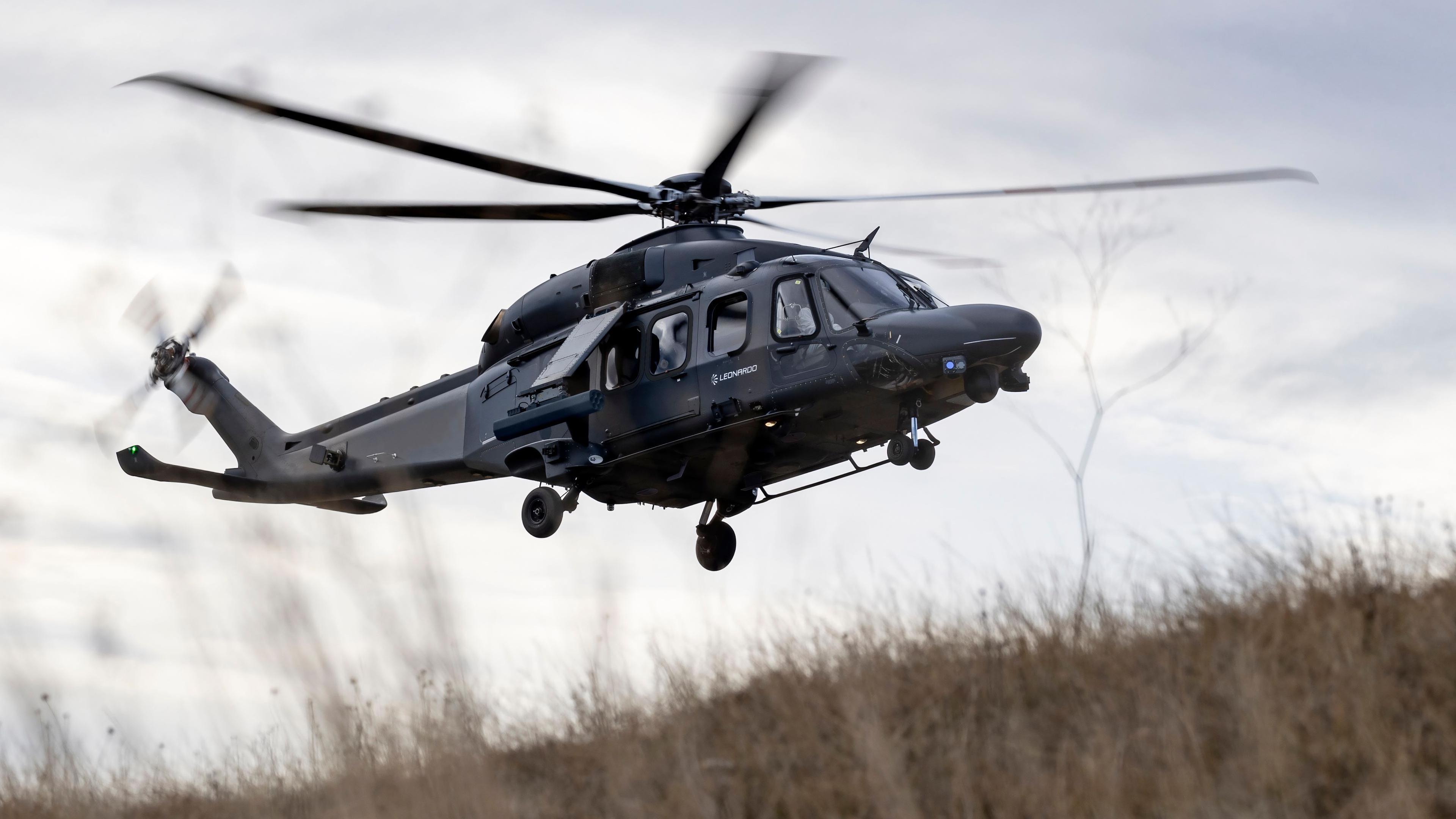 A helicopter flying. It is black. The camera is looking up. Grass on the floor is blowing from the rotors.