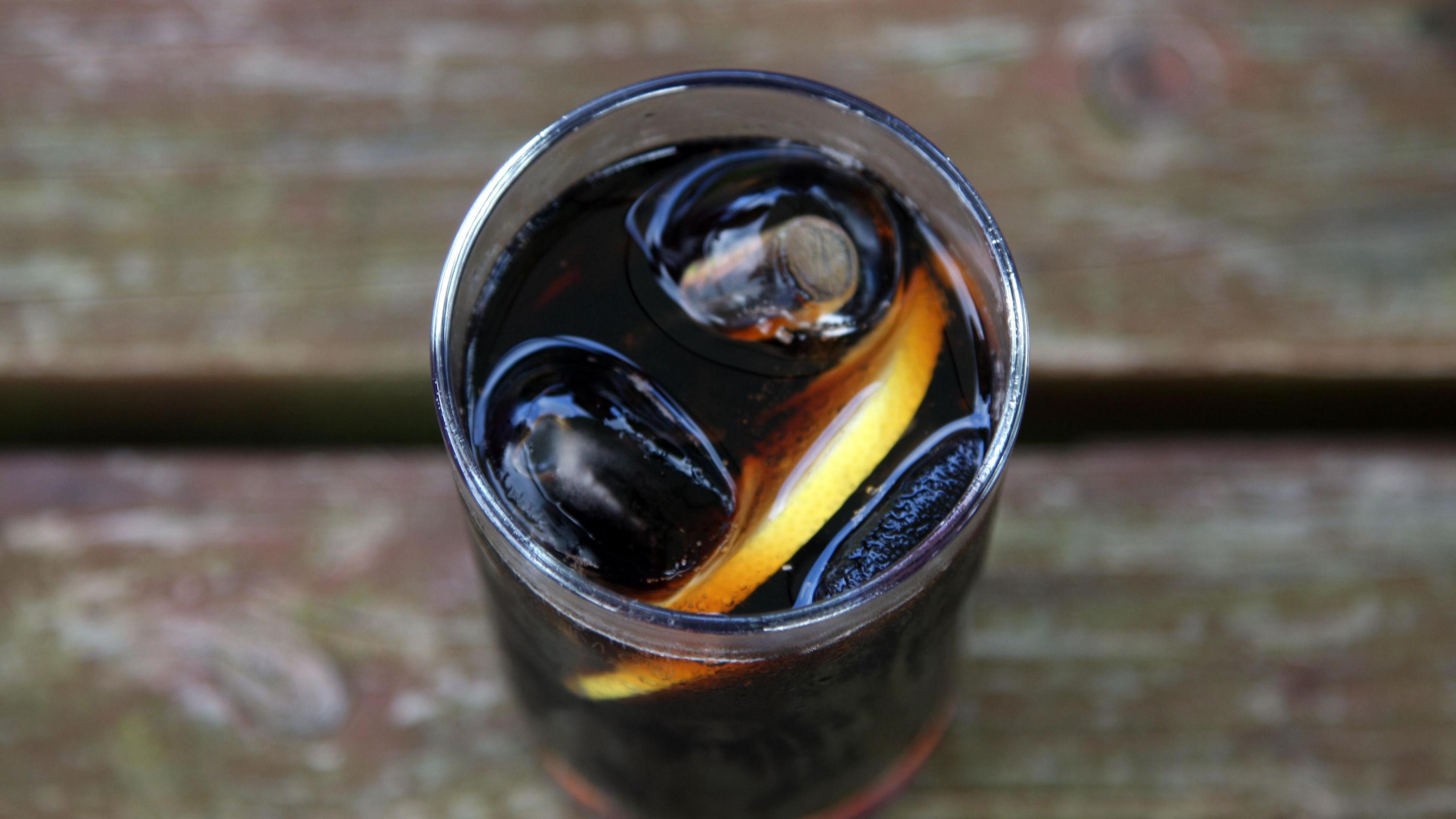 A glass drink's tumbler with dark liquid in it, ice cubes and a lemon slice. It is on a wooden table. 