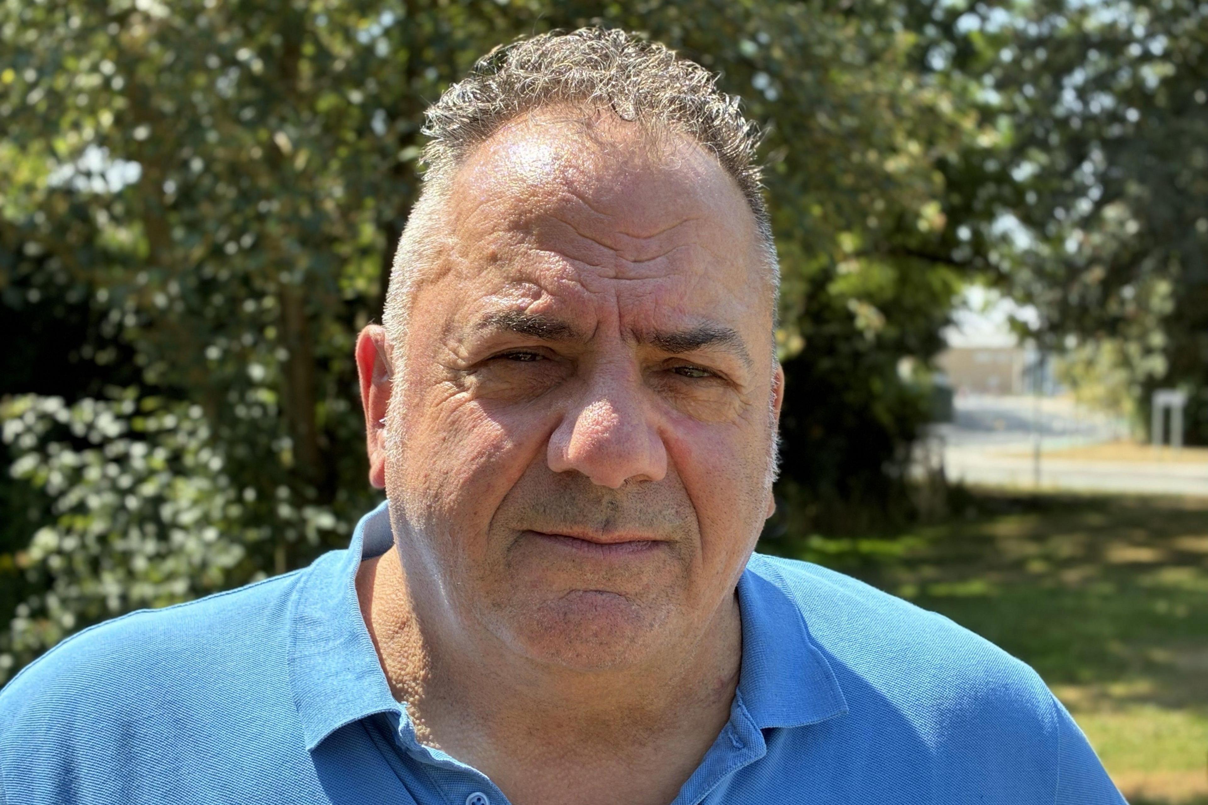 A middle-aged man with short cropped hair, curly on top, stands in sunlight in  front of a patch of grass and green trees. He is wearing a blue polo shirt and has a serious expression on his face.