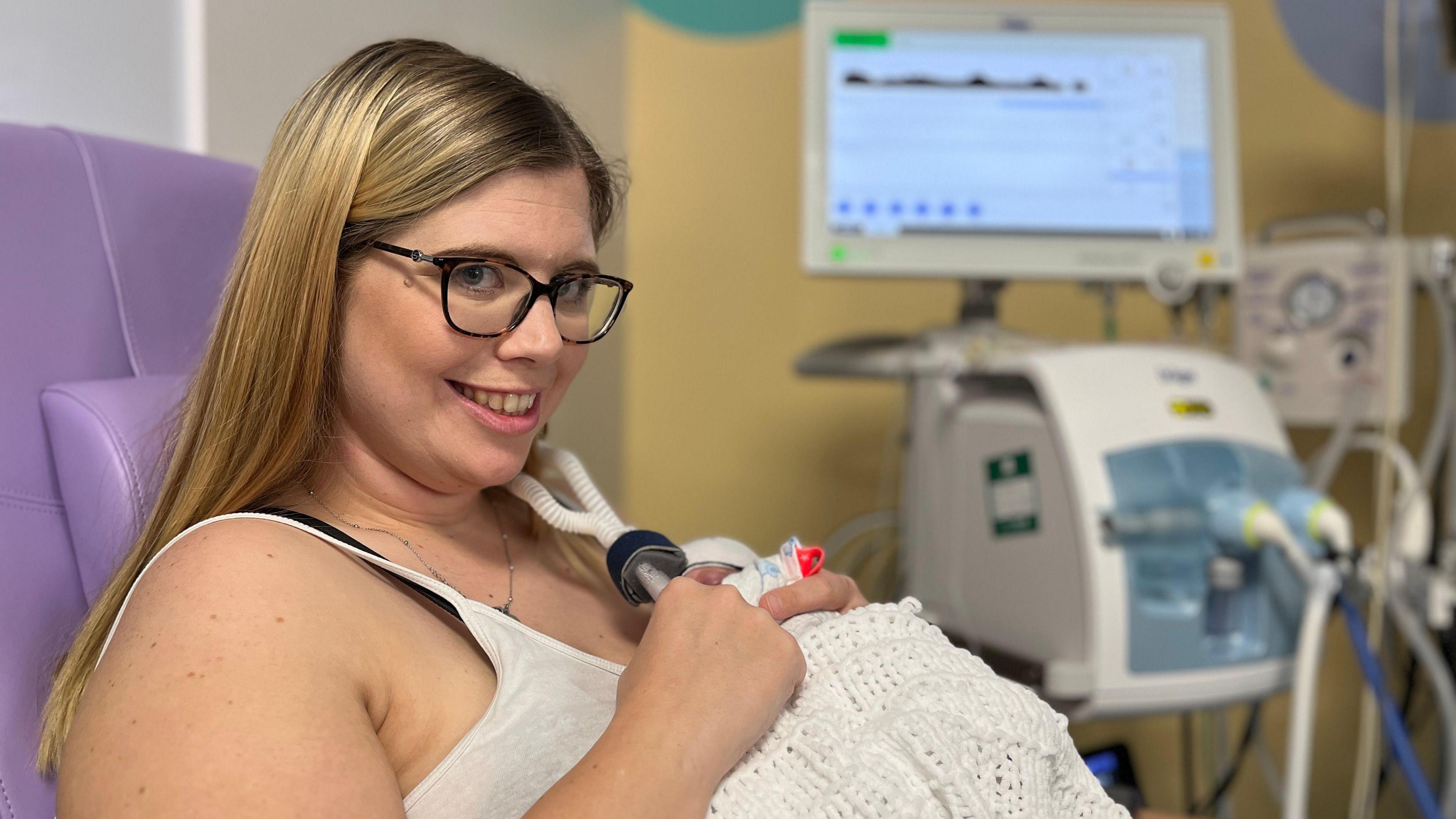 Lauren has long blond hair and wears glasses. She is sitting in a purple hospital chair, holding her baby Zachary in a blanket on her chest. Zachary is attached to a ventilator machine, which can be seen in the background.