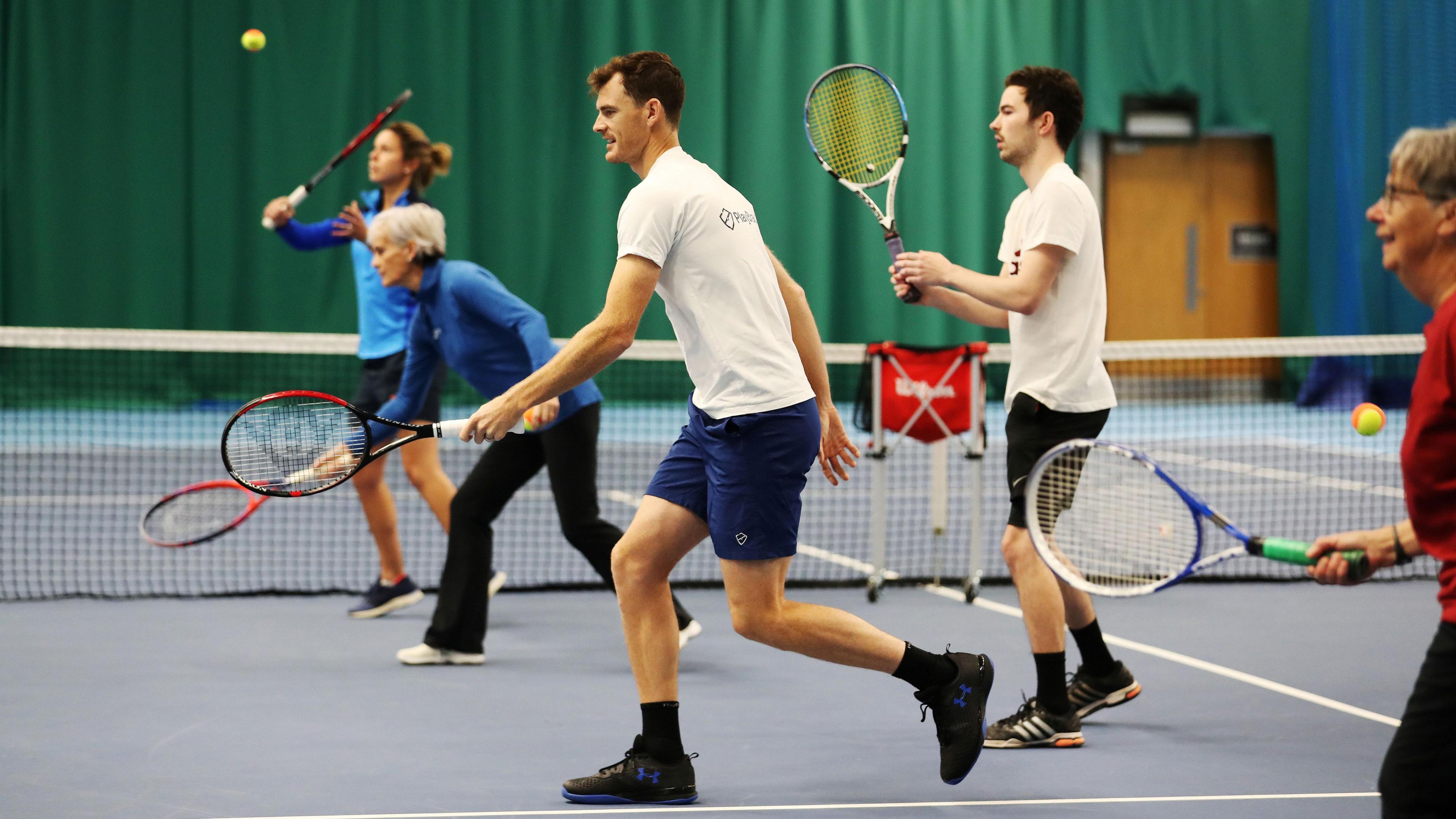 A row of people playing tennis indoors. They are all holding racquets and hitting various balls.