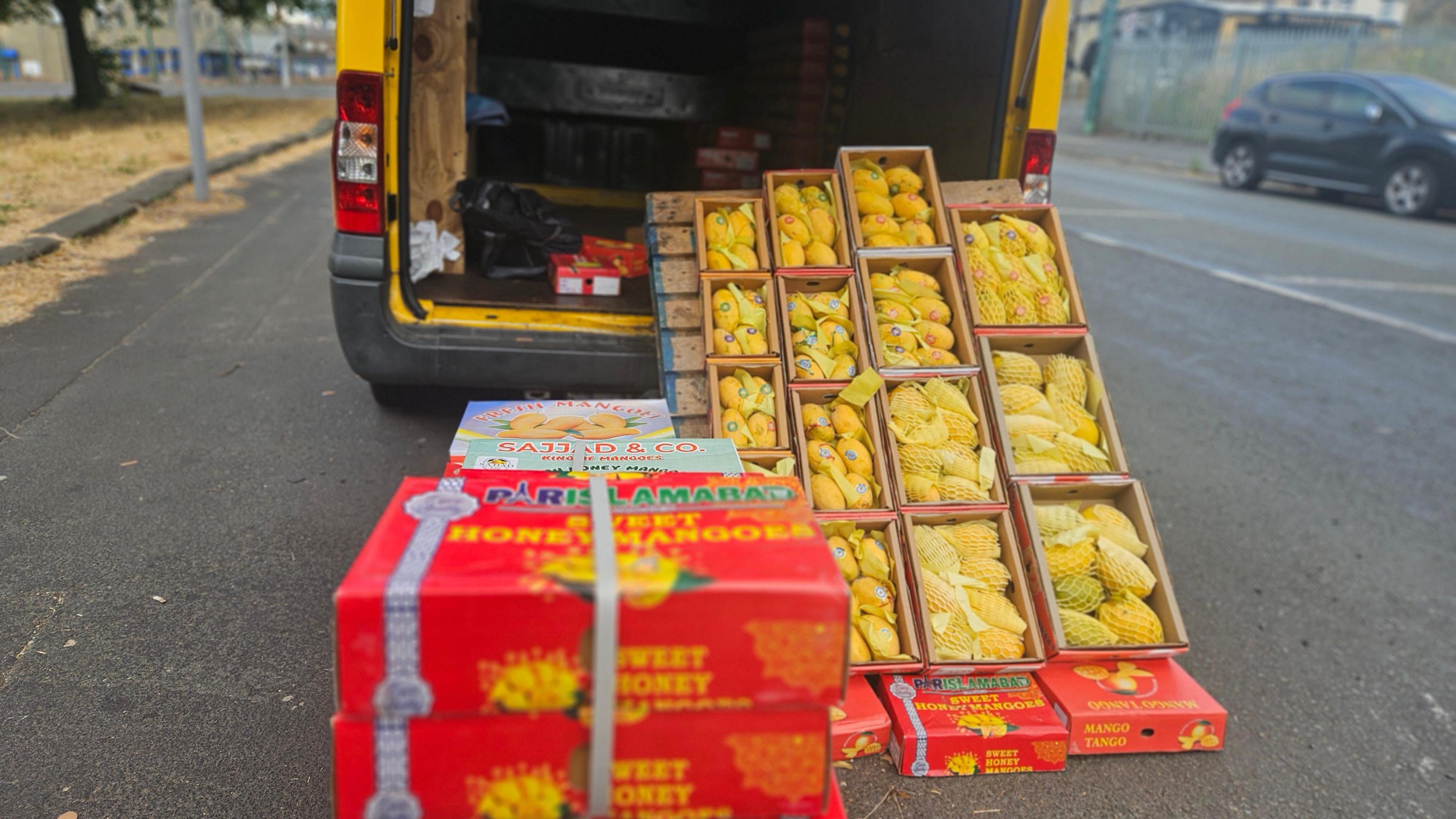 A yellow van parked on the side of a road. Piled up outside it, and visible inside, are multiple small crates of bright, ripe mangoes. The fruits are strapped together in closed boxes, or are on display in open boxes with a soft protective netting covering them.