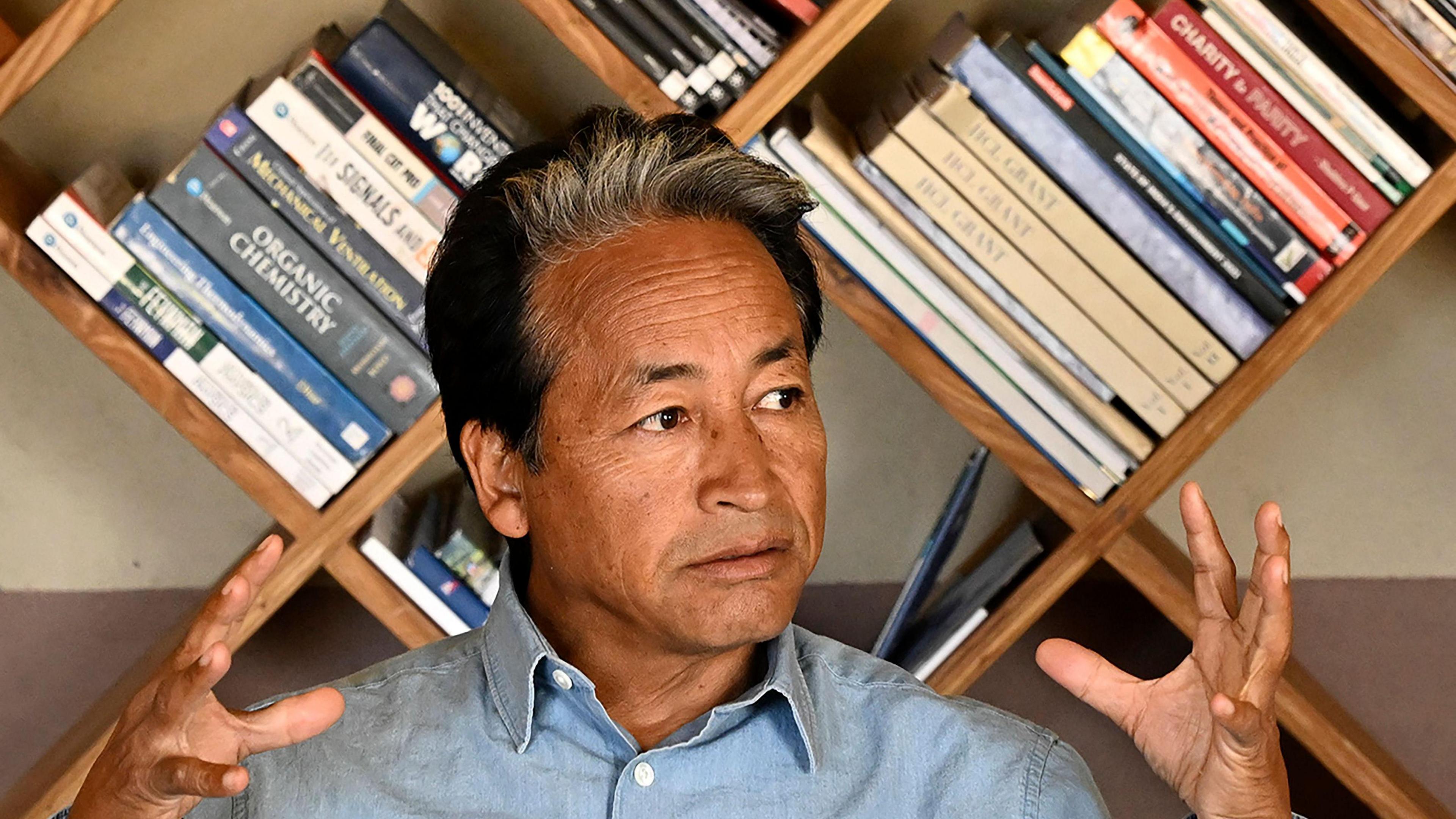 Sonam Wangchuk, wearing a blue shirt, sits in front of a book shelf, and looks away from the camera 