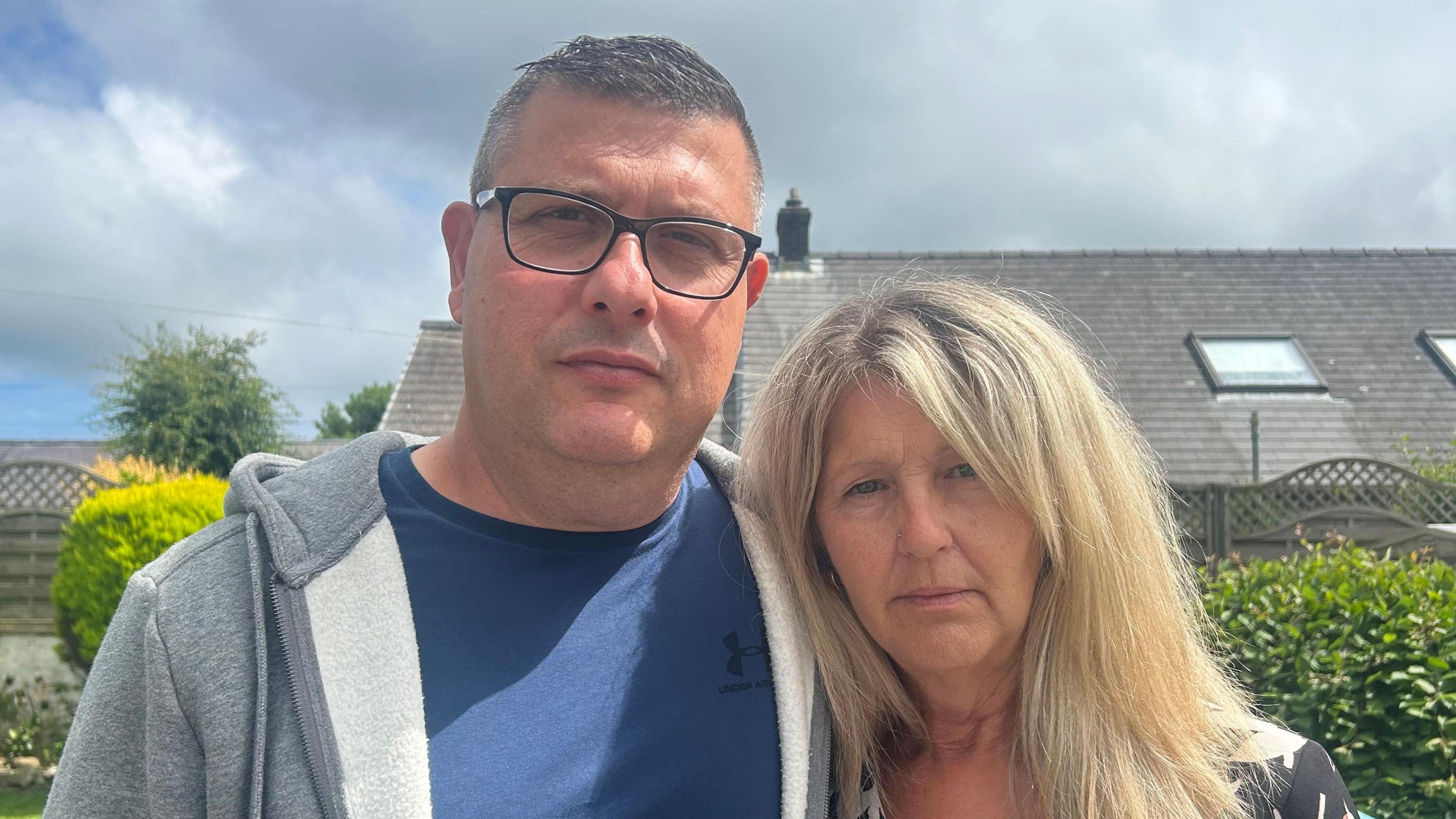 Brian Buckle (chwith) - dyn gwallt byr llwyd mewn crys-t glas a hoodie llwyd - gyda'i wraig, Elaine - menyw gwallt hir melyn mewn top blodeuog du, coch a gwyn - y ddau yn sefyll yn yr ardd ac yn edrych tua'r camera