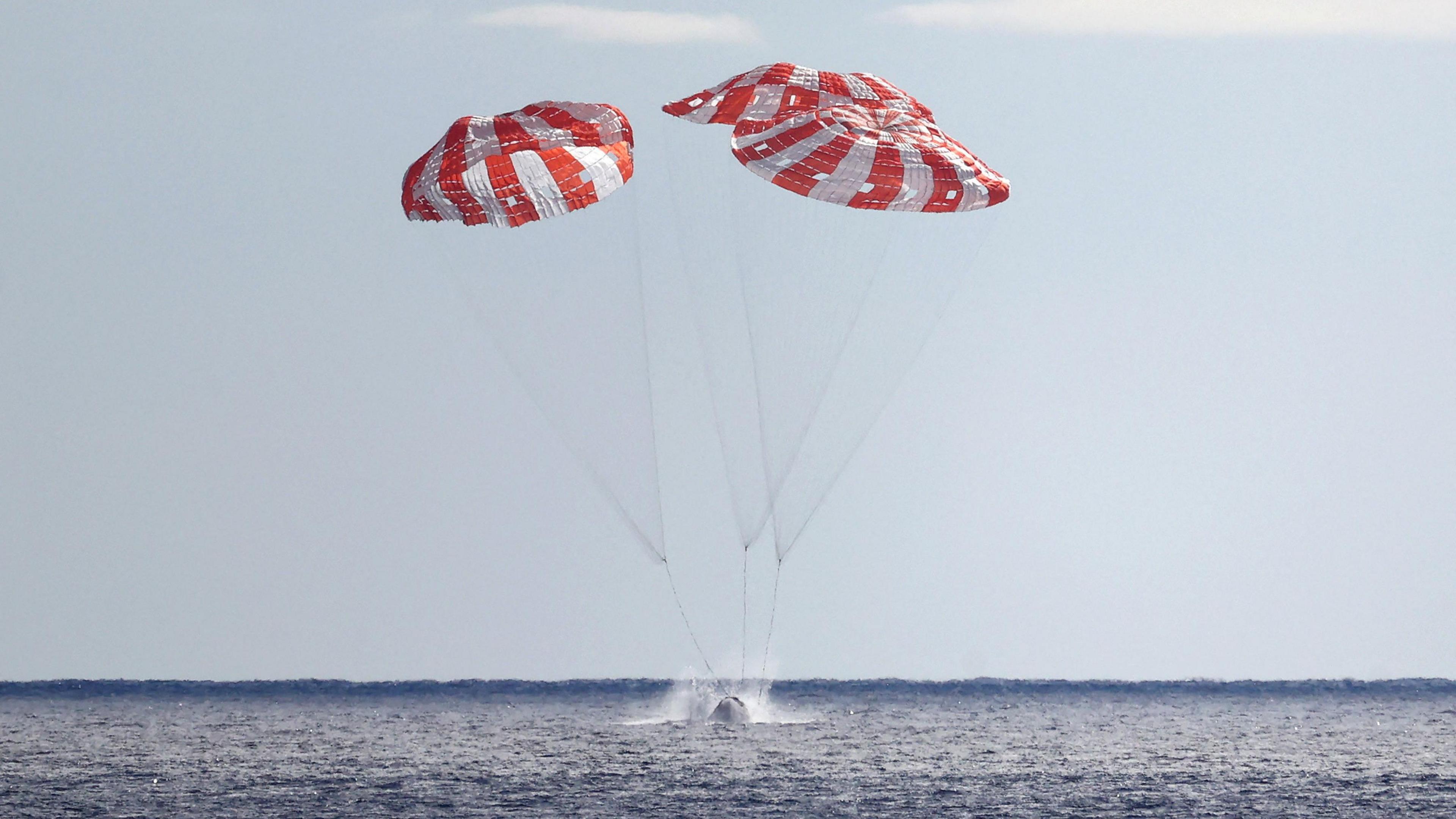 red and white parachutes above a capsule that's splashing into the sea