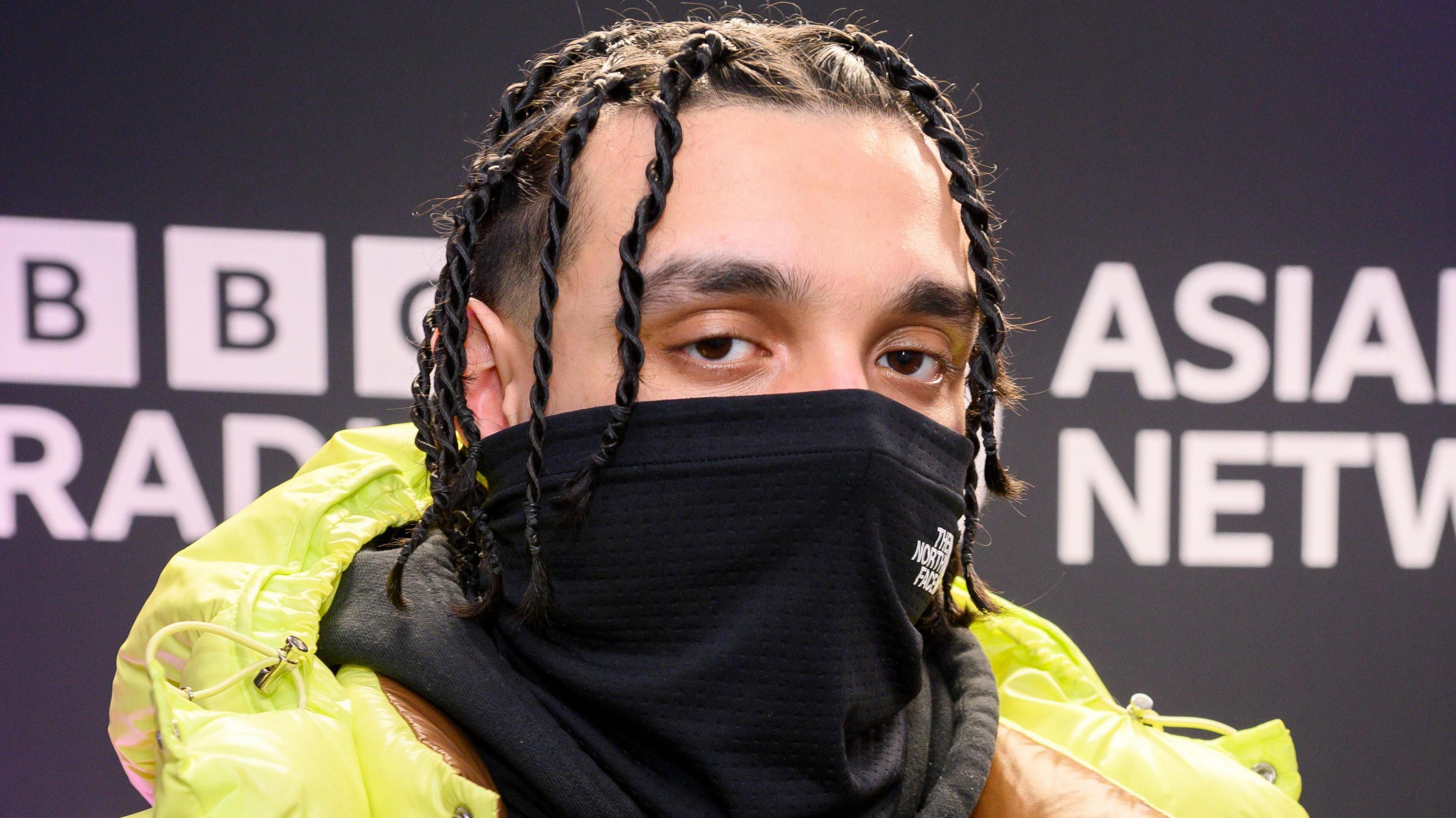 Sliime in the BBC Asian Network studio in February 2024. The rapper wears a bright green puffer jacket and covers the bottom half of his face with a black scarf. 