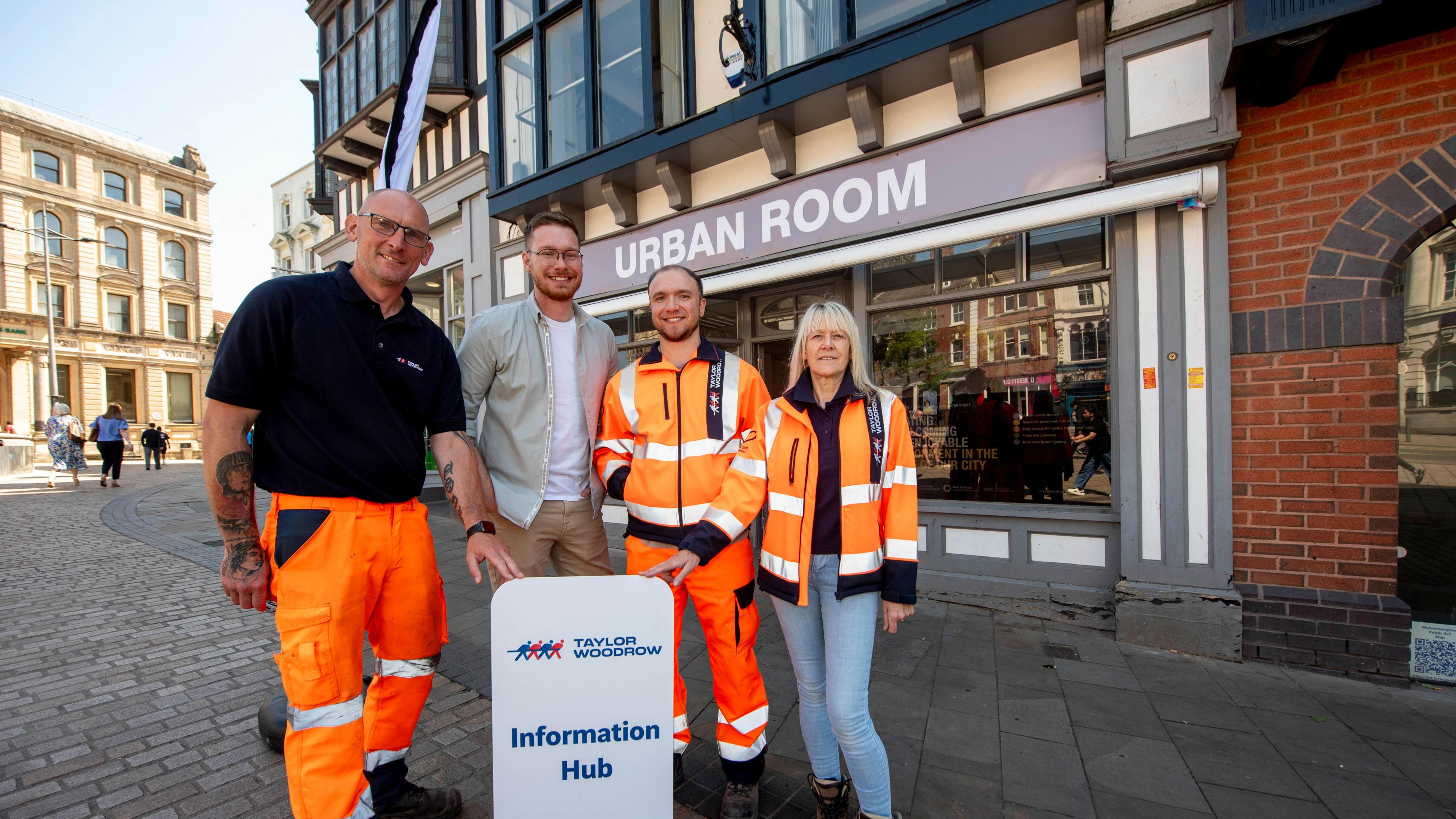 A woman and three men stand together outside the Urban Room in Wolverhampton city centre. Two of them touch a white sign saying 'Taylor Woodrow Information Hub' on it. The woman wears an orange hi-vis jacket and two of the men also wear hi-vis clothing. The other man is in a light coloured shirt with a white T-shirt underneath and brown trousers.