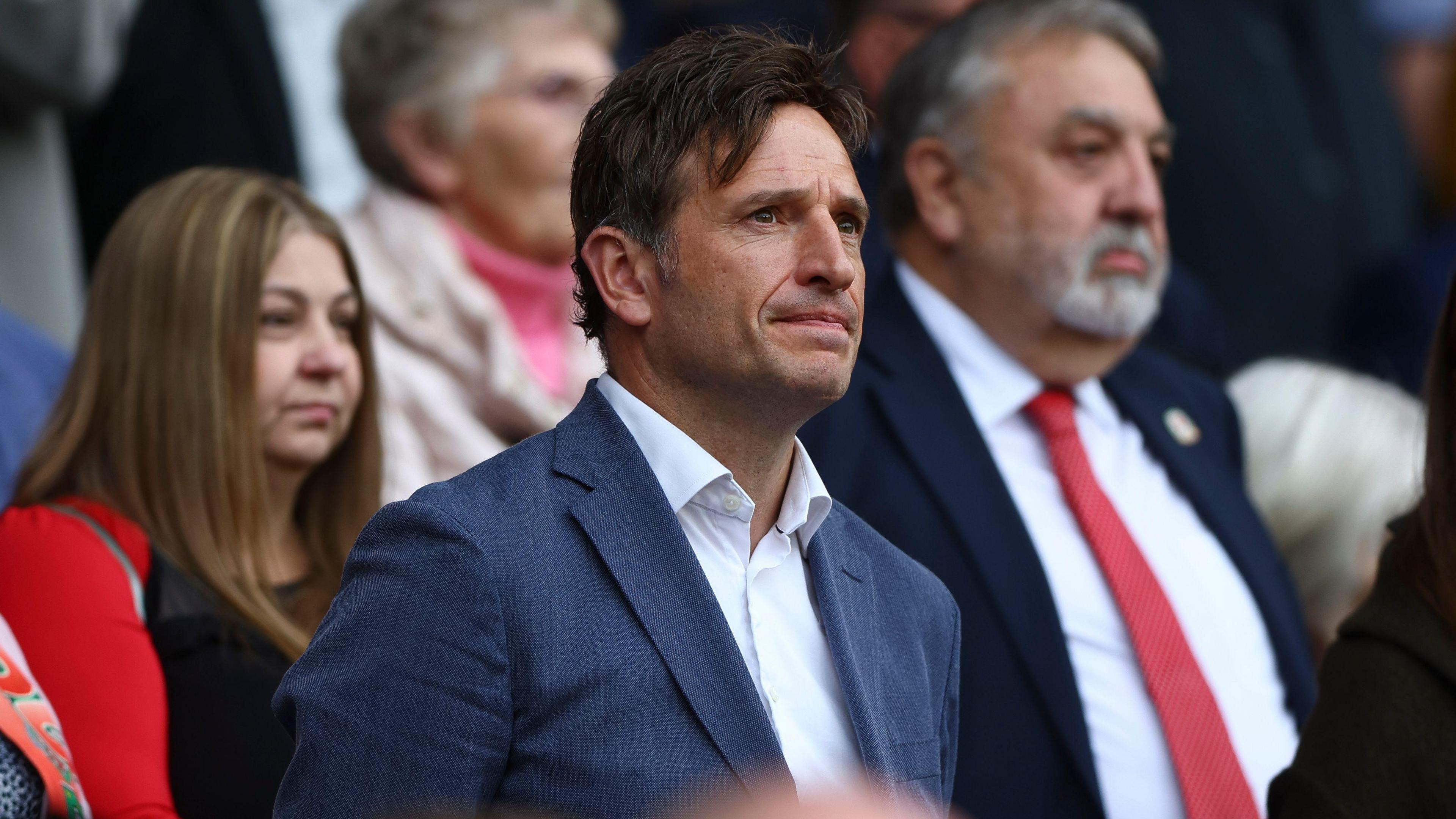 FAW chief executive Noel Mooney, in a blue suit and open neck white shirt, stands during a game.