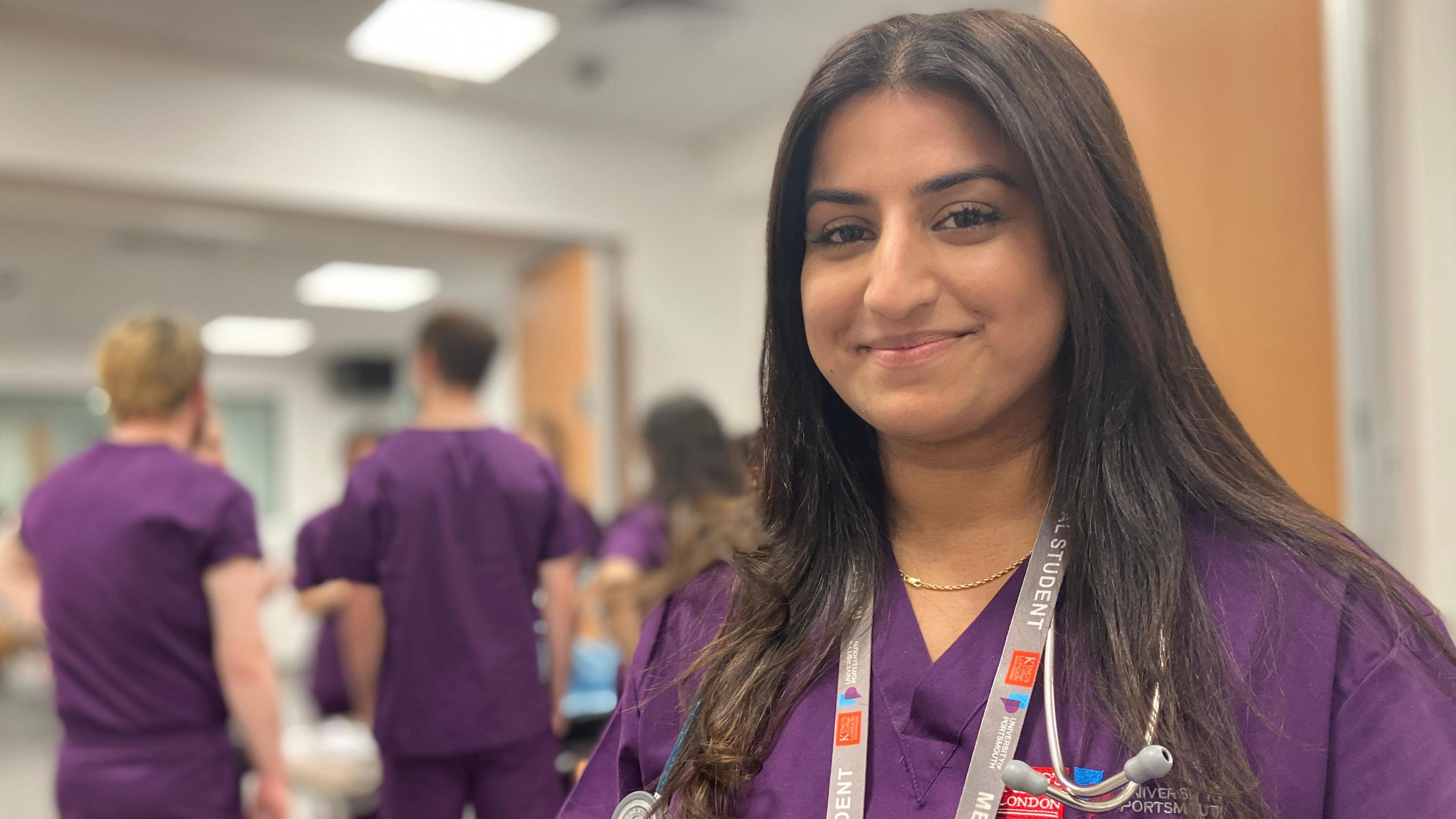 Head and shoulders of Kareena Panesar in purple medical clothing, with other medical students in the background