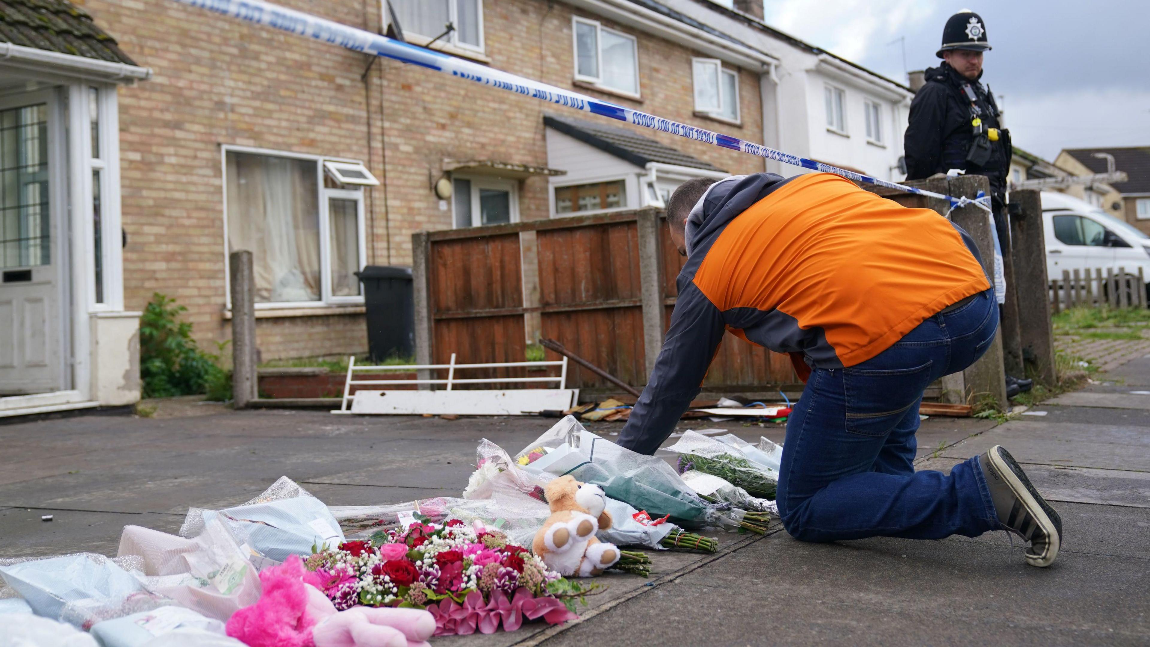 A man is laying down a bunch of flowers in front of a house. There is police tape above him and more flowers and cuddly toys can be seen on the ground 