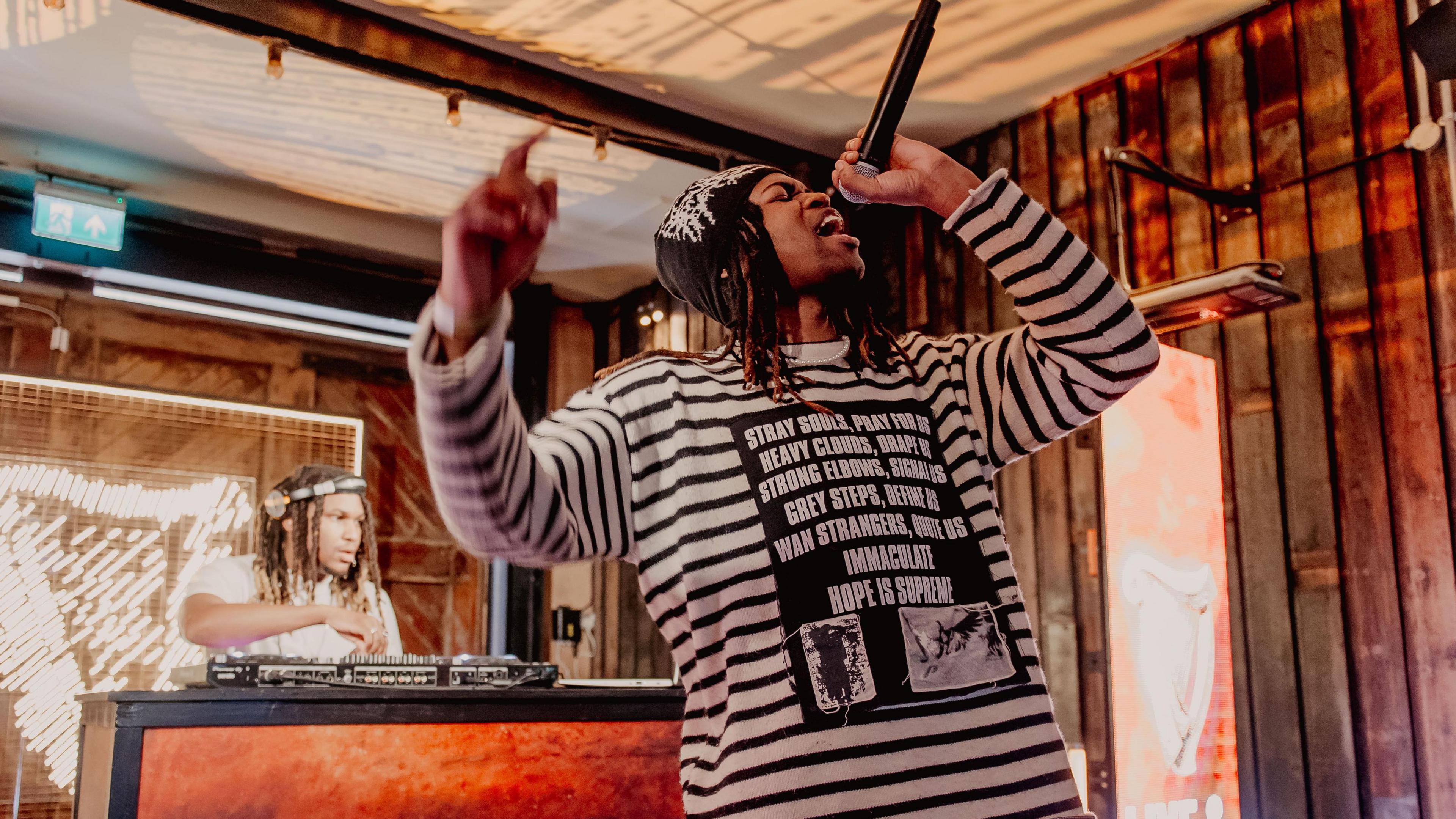 Jordan Adetunji performing onstage. He's wearing a stripped top and black trousers and holding the mic over his head. A dj can be seen behind him.