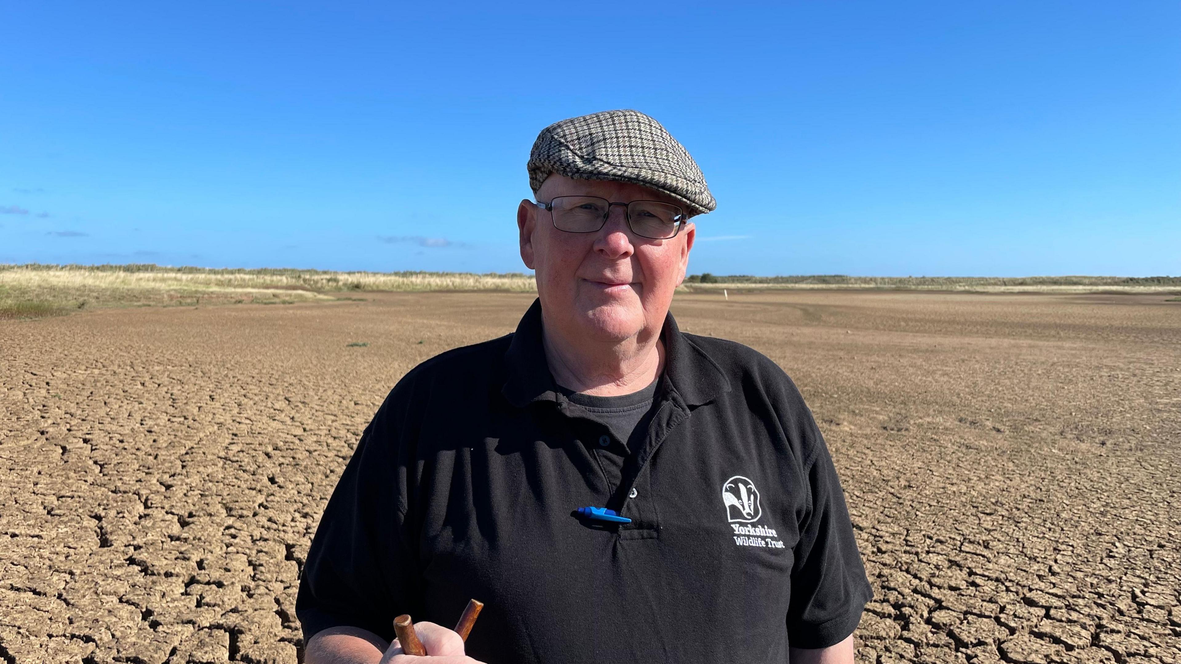 A man holding a walking staff and wearing a flat cap is standing in a dried-up terrain.