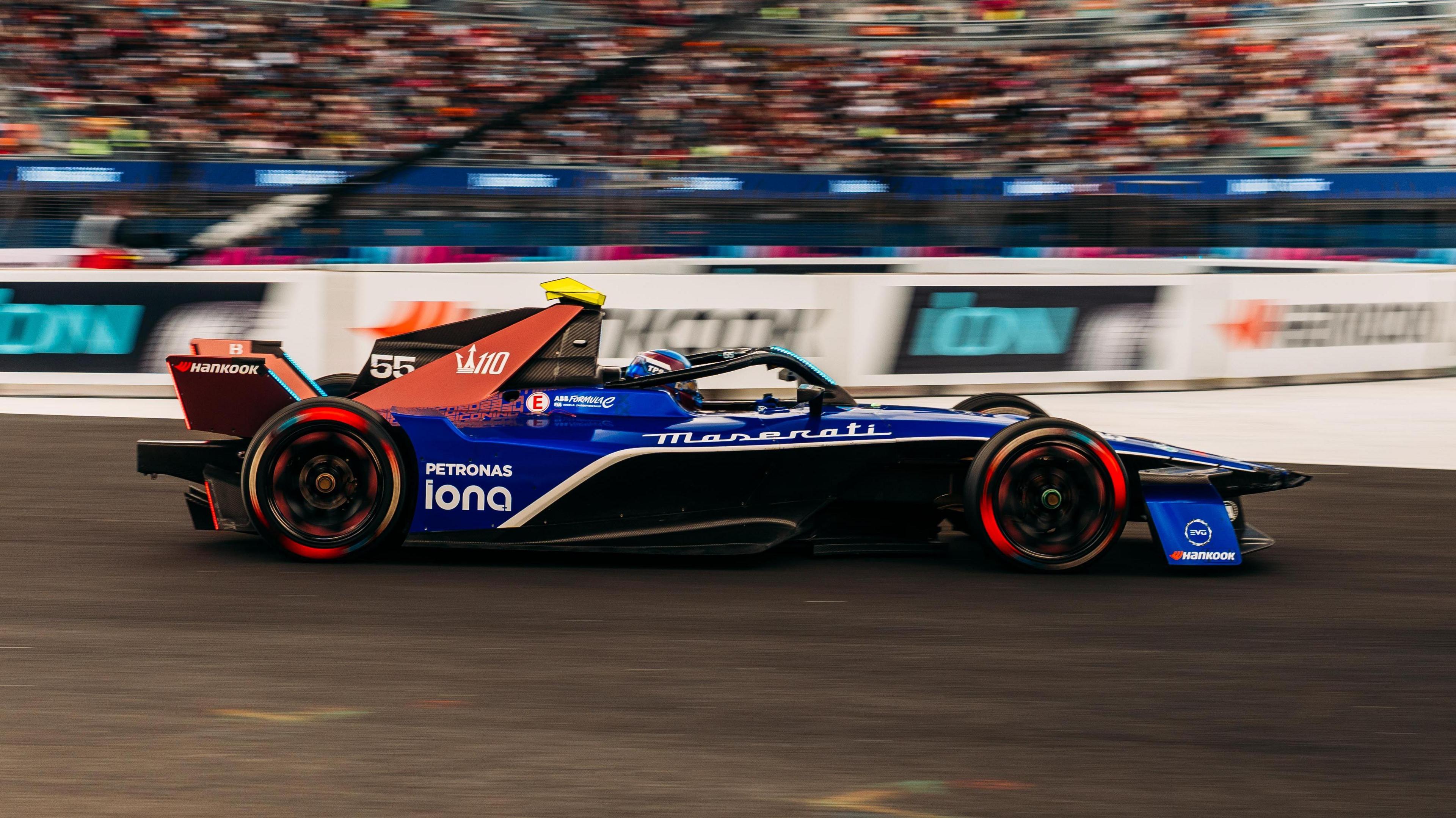 A blue and red racing car on a racing track with the audience and advertising boards blurred in the background.