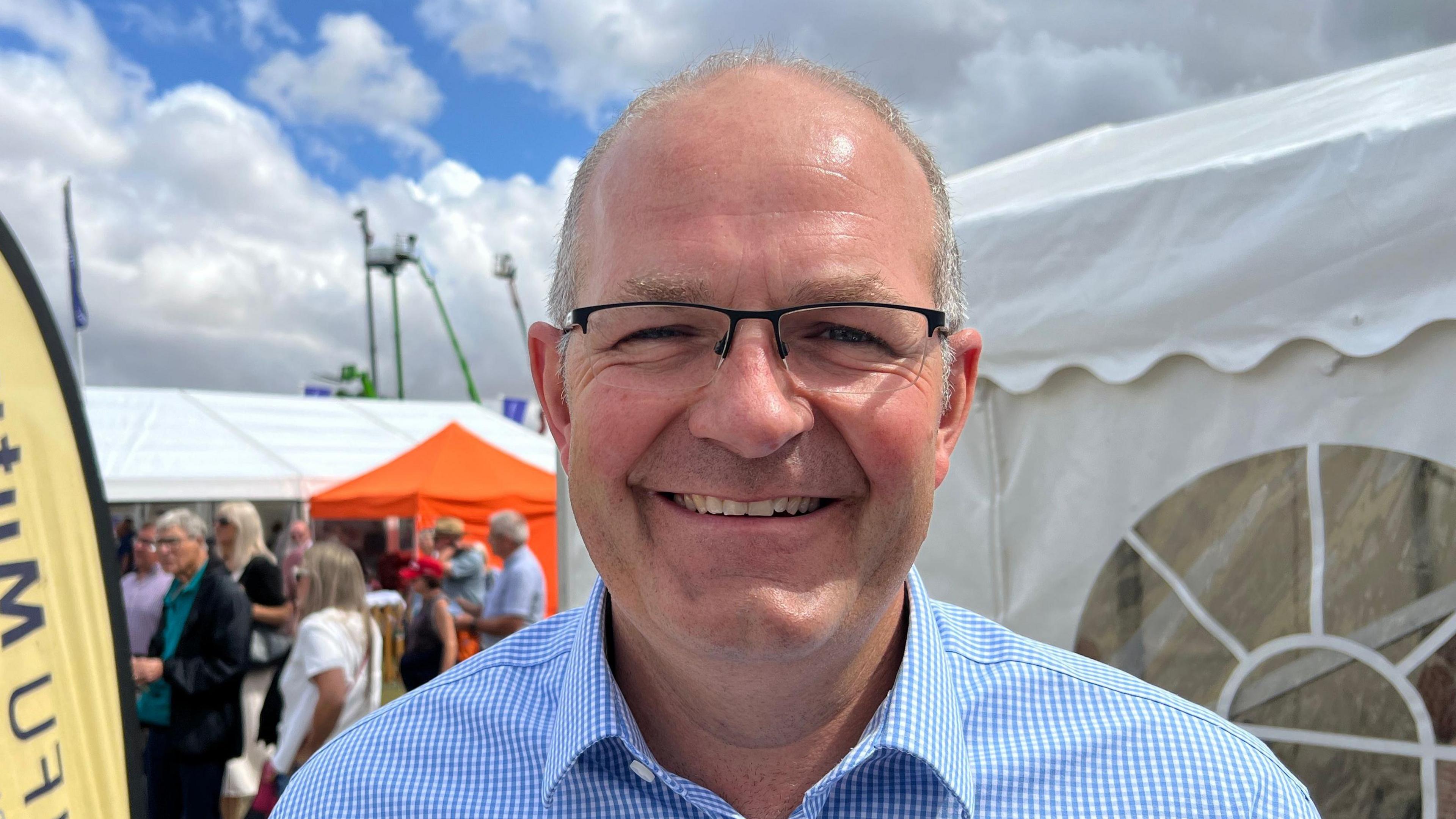 The photo shows a man smiling at the camera. He is wearing glasses, and has a blue and white checked shirt on. He's standing in front of a marquee, and there is a crowd of people behind him. There are also cranes in the distance.