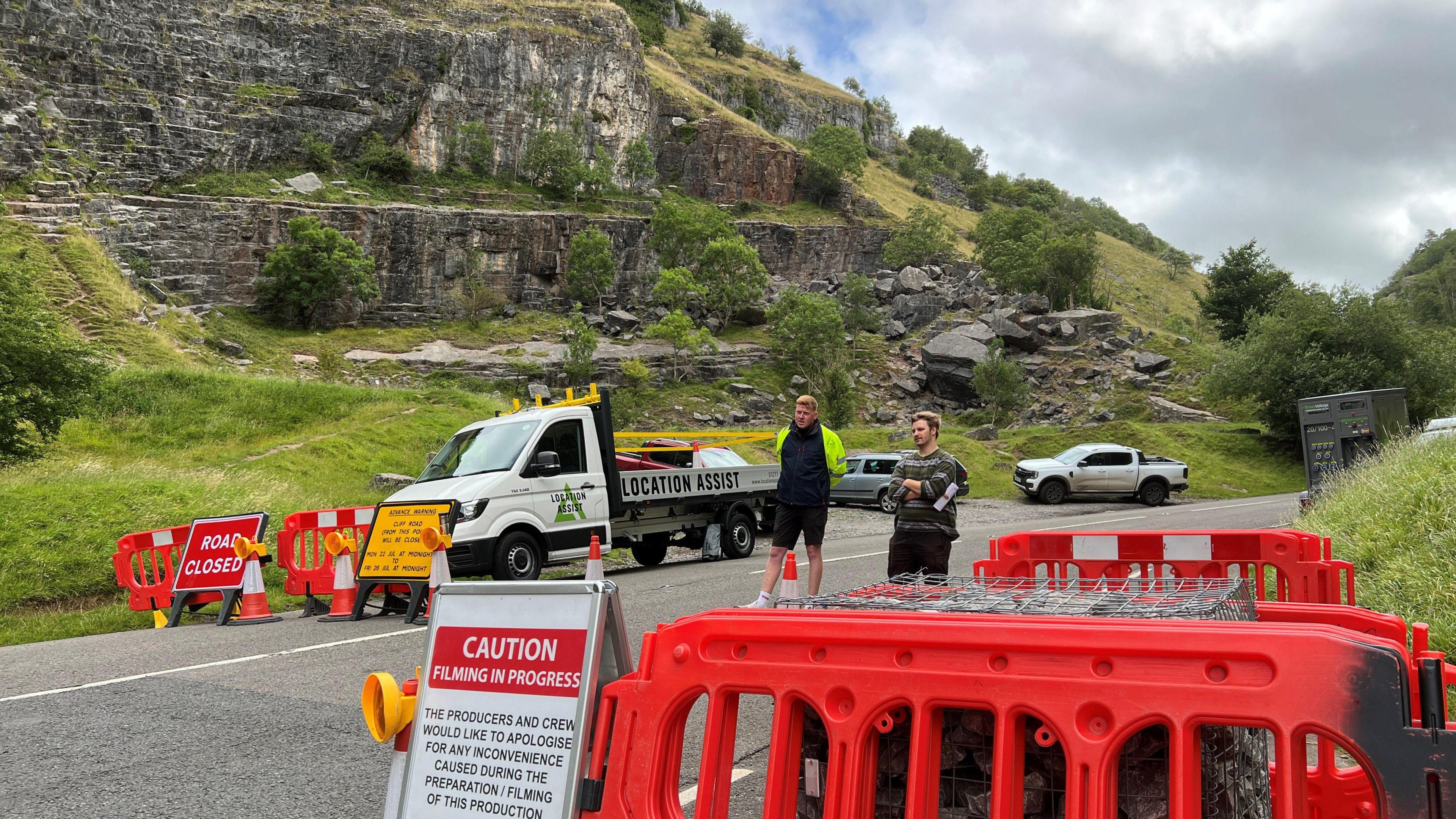 Cheddar Gorge cliffs with road closed sign and red barrier and people walking down street. 