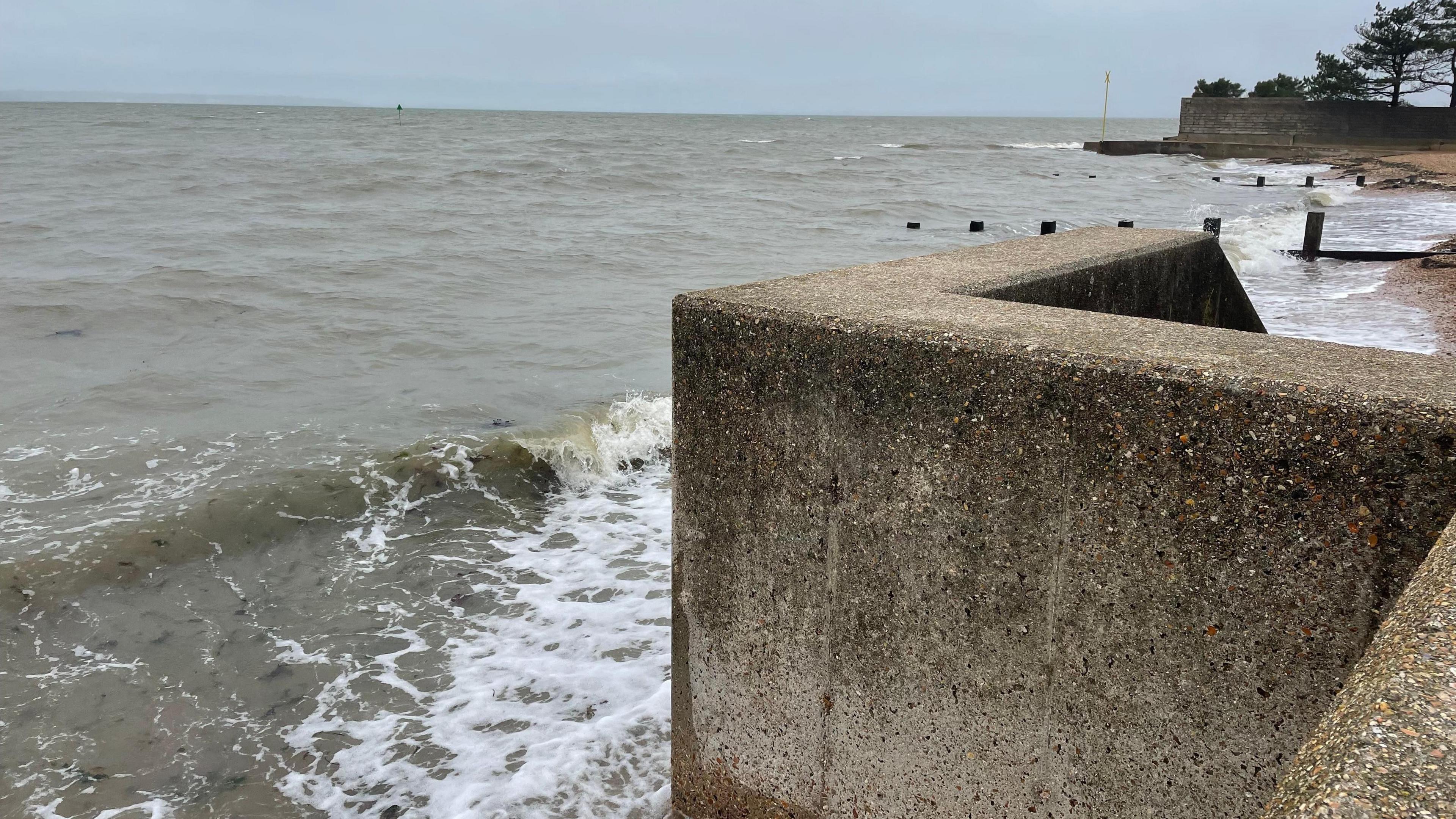 A concrete seawall overlooks the sea which is frothy. It looks like strong winds have been swelling the waves under grey skies. There is no-one in the picture