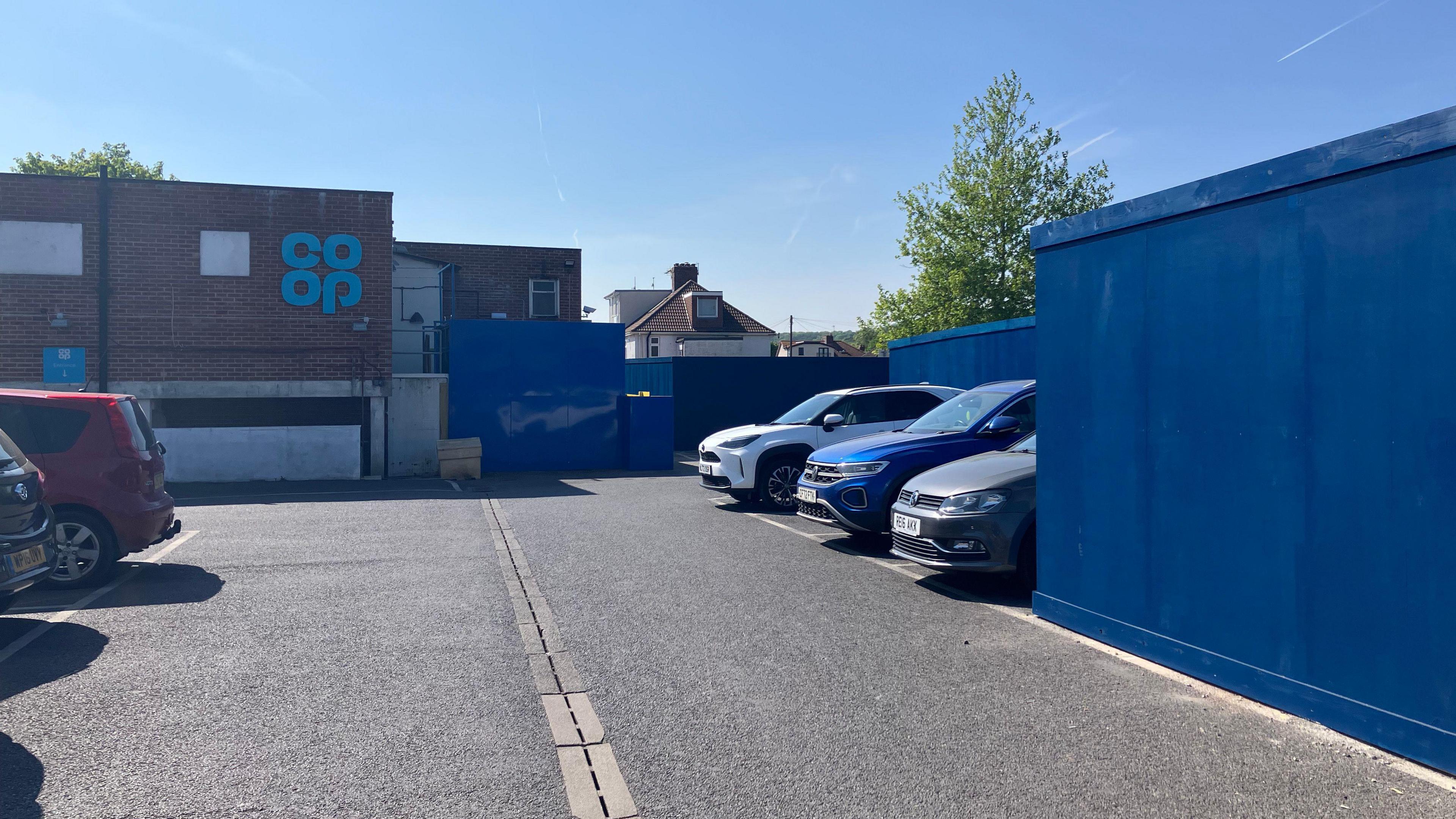 Large wooden walls painted blue installed in a car park. There are cars parked in spaces, and a sign for Co-op in the distance.