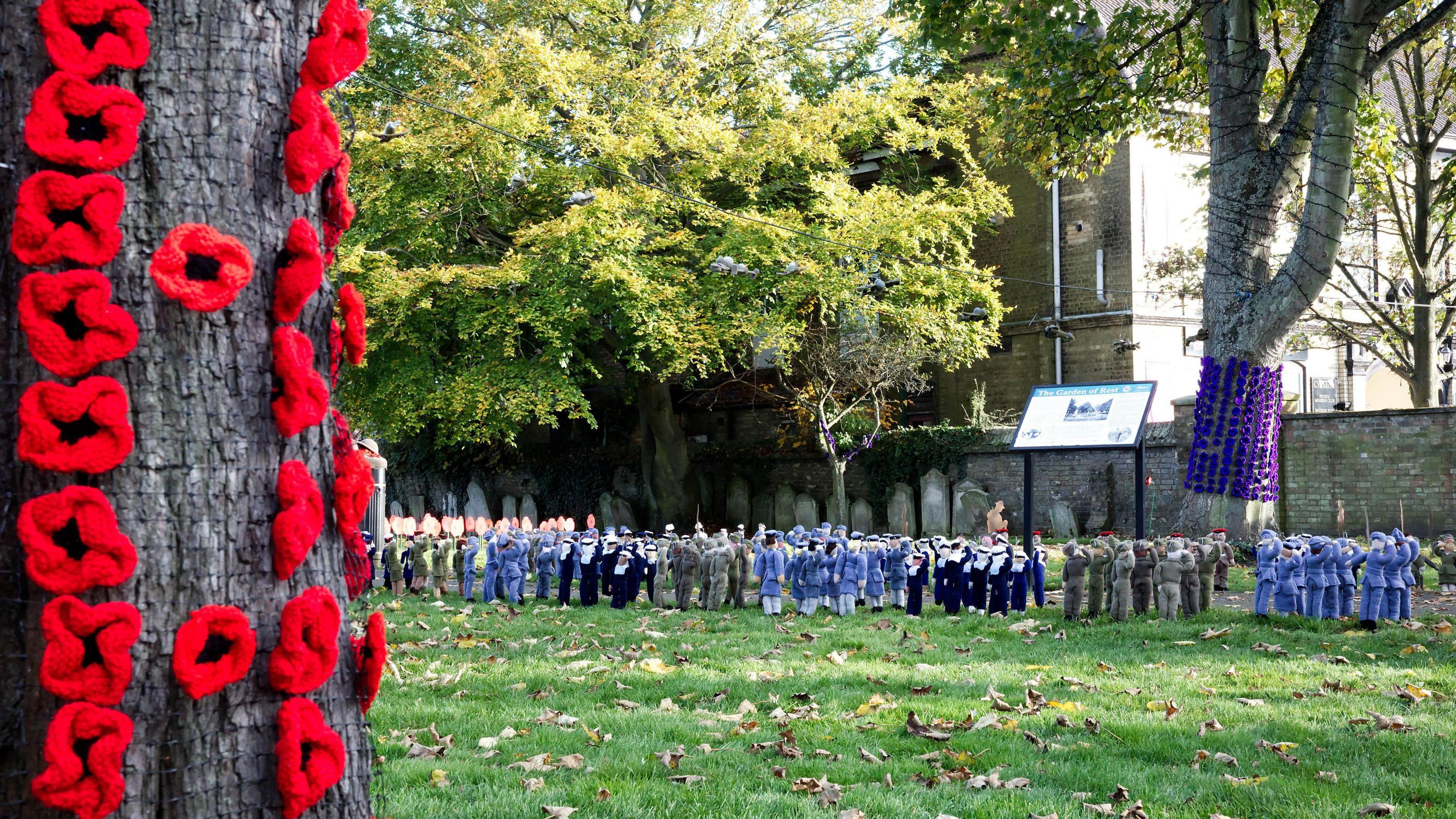 Tree bark covered with red knitted poppies with knitted small soldiers placed on the grass in the distance