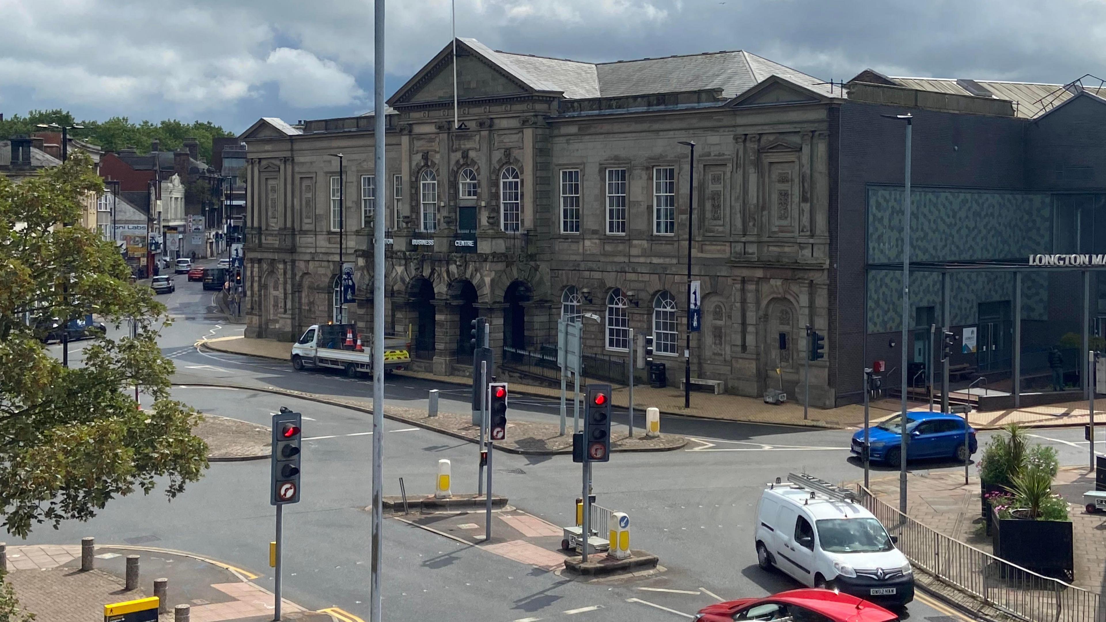 Times Square, Longton, has a traffic light junction near the town hall, where several roads meet. Lights are red in the photograph and cars are using the junction. There is street furniture with bollards and trees.