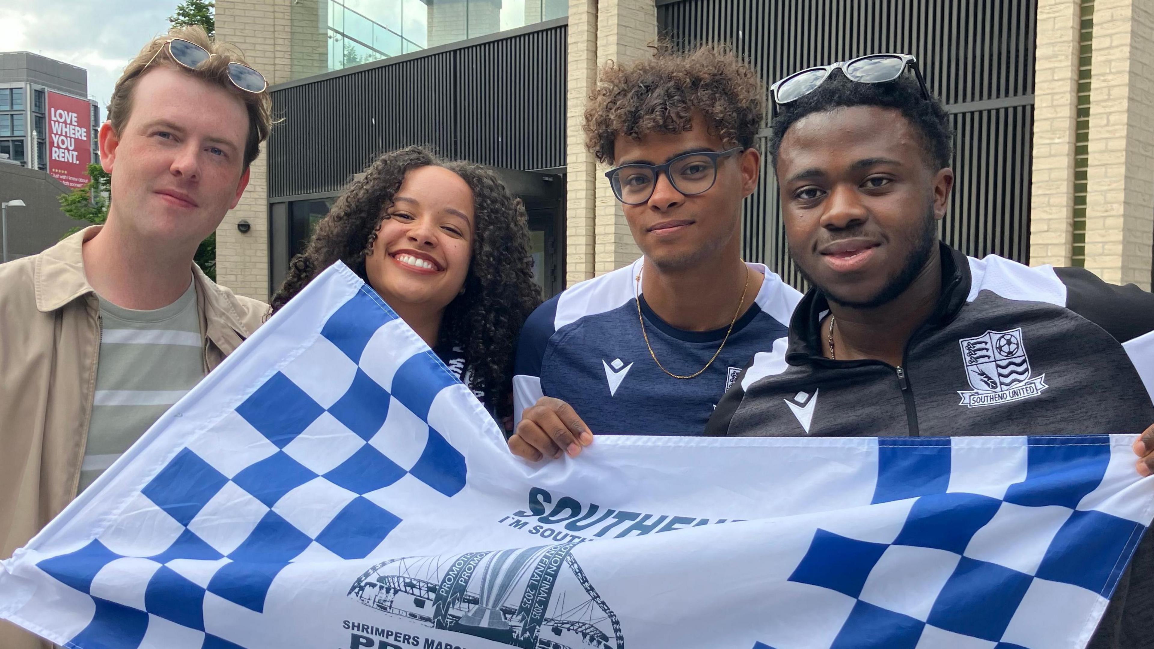Niamh Bligh, who has curly brown hair, smiling at the camera. She is flanked by three men. They are all holding a blue and white chequered Southend flag.