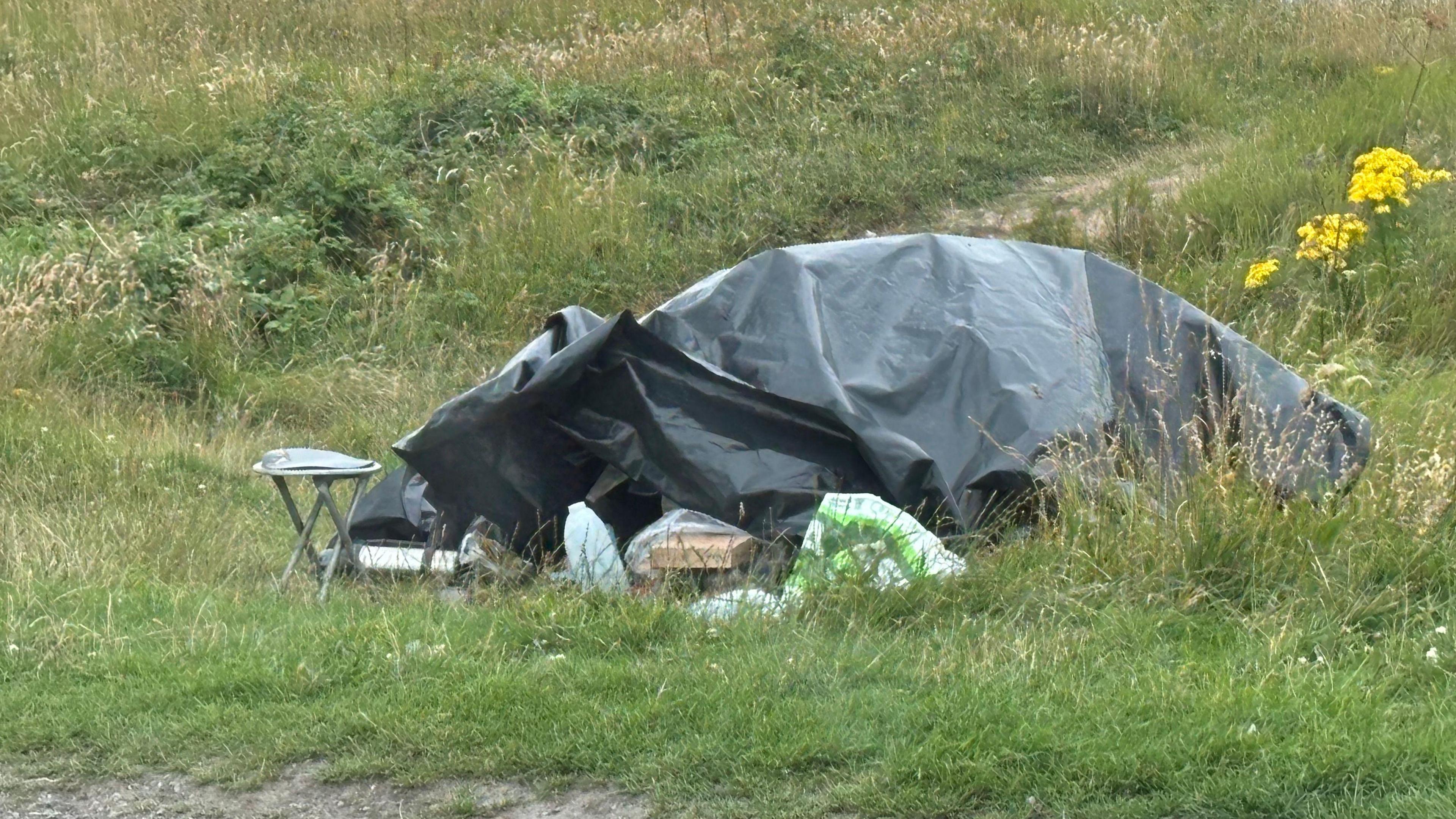 A vehicle covered with tarpaulin and secured by bricks with a stool left outside. 