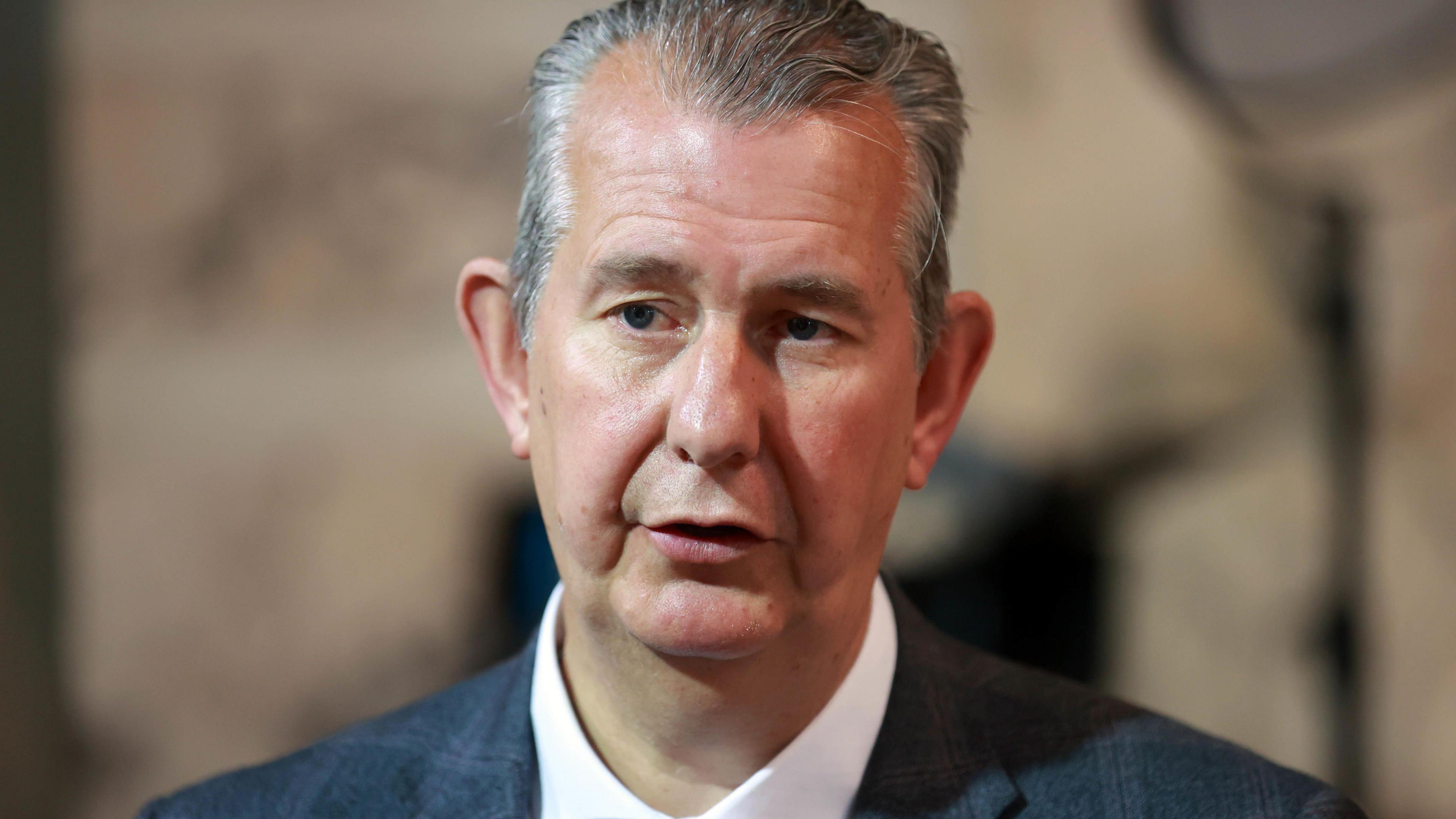 A close-up image of Edwin Poots. He is an older man with short grey hair. He is wearing a dark plaid suit jacket and a white shirt. He is speaking into a microphone but the background is blurred.