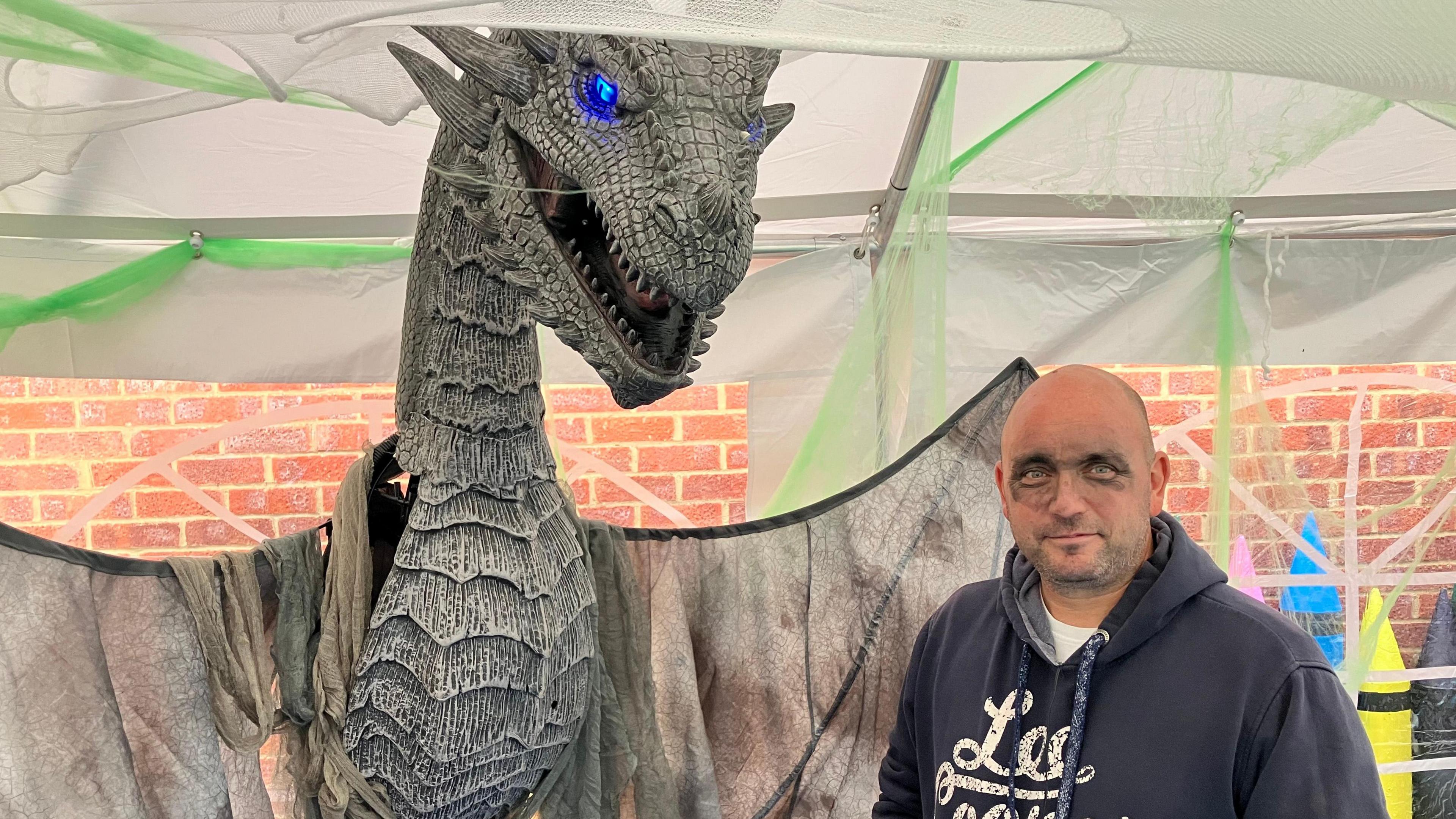 A man in a black hoodie with some black face paint stands next to a large animatronic dragon.