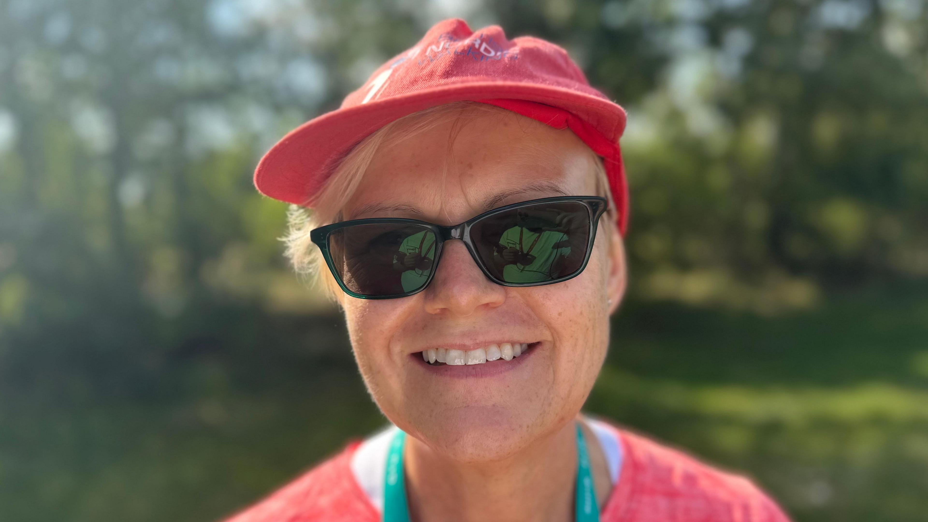 A smiling Anne Mills wearing a hat and sunglasses looks straight at the camera.