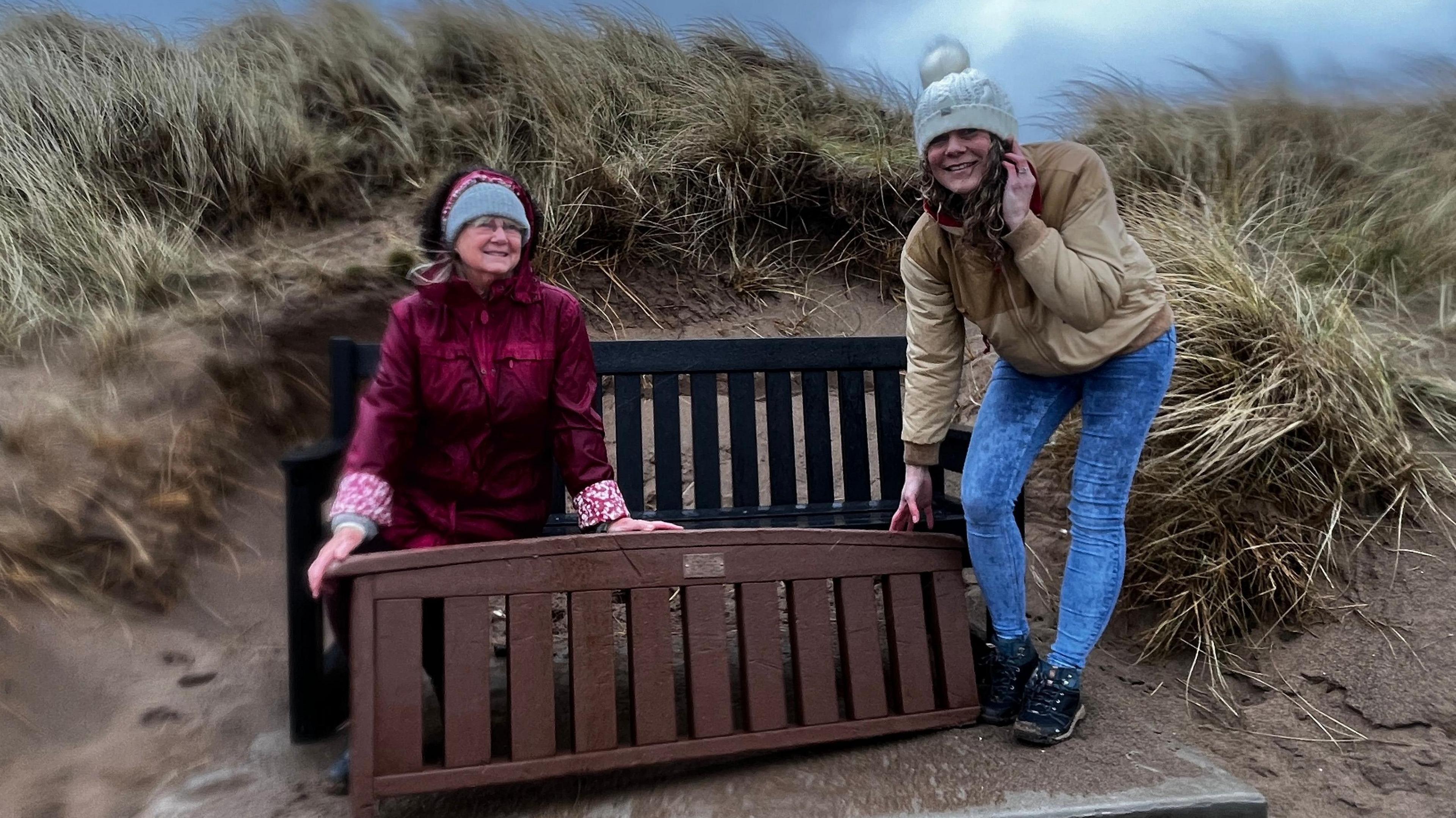 Two women hold up the backrest of the newly-rediscovered memorial bench while sat on another bench on a beach. Marram grass can be seen all around them blowing in the wind. 