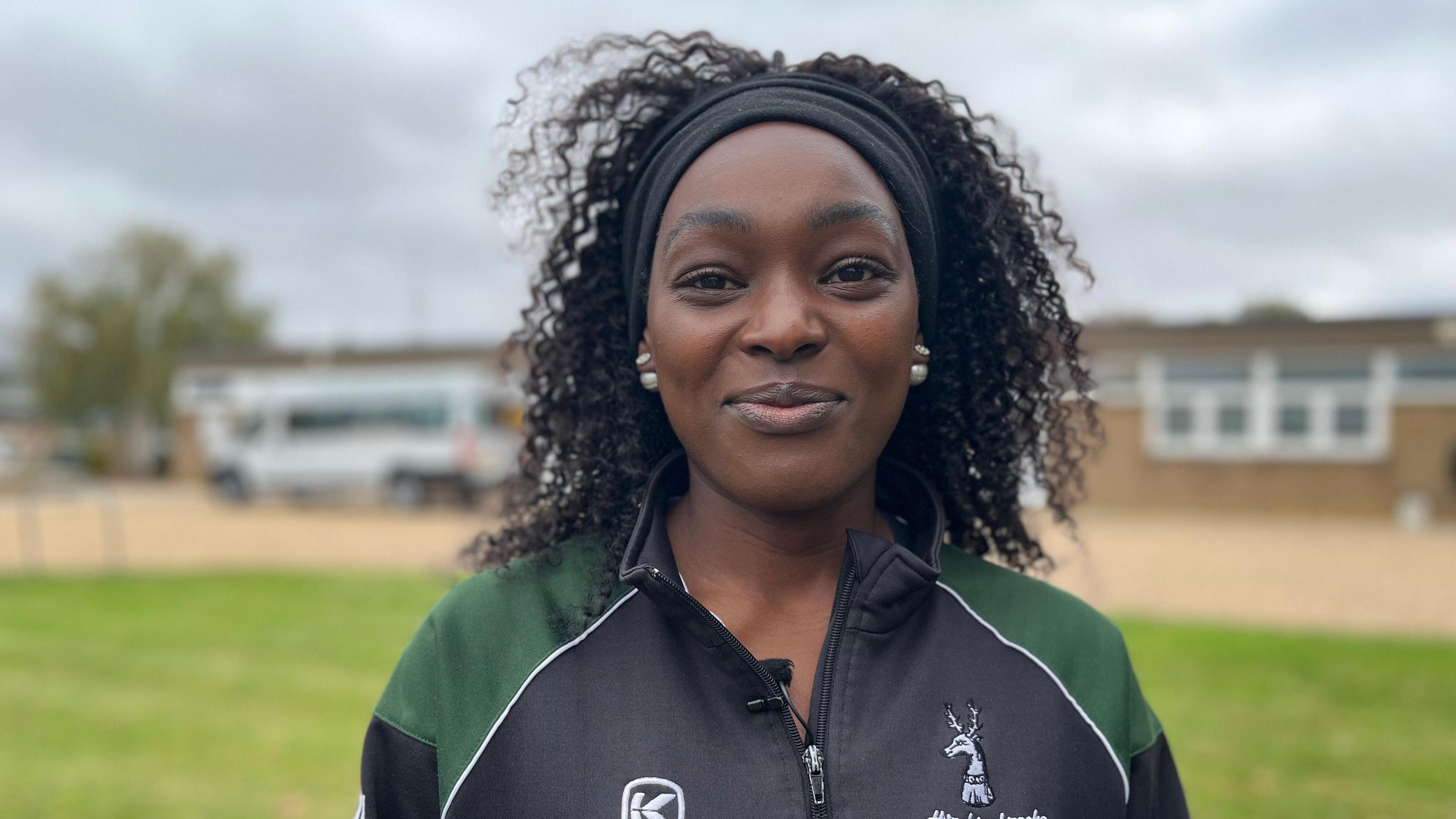 Femi Solano is looking at the camera with a closed mouth smile. She has dark skin and dark hair with a black hairband keeping her hair off her face. She wears a green and black tracksuit top.