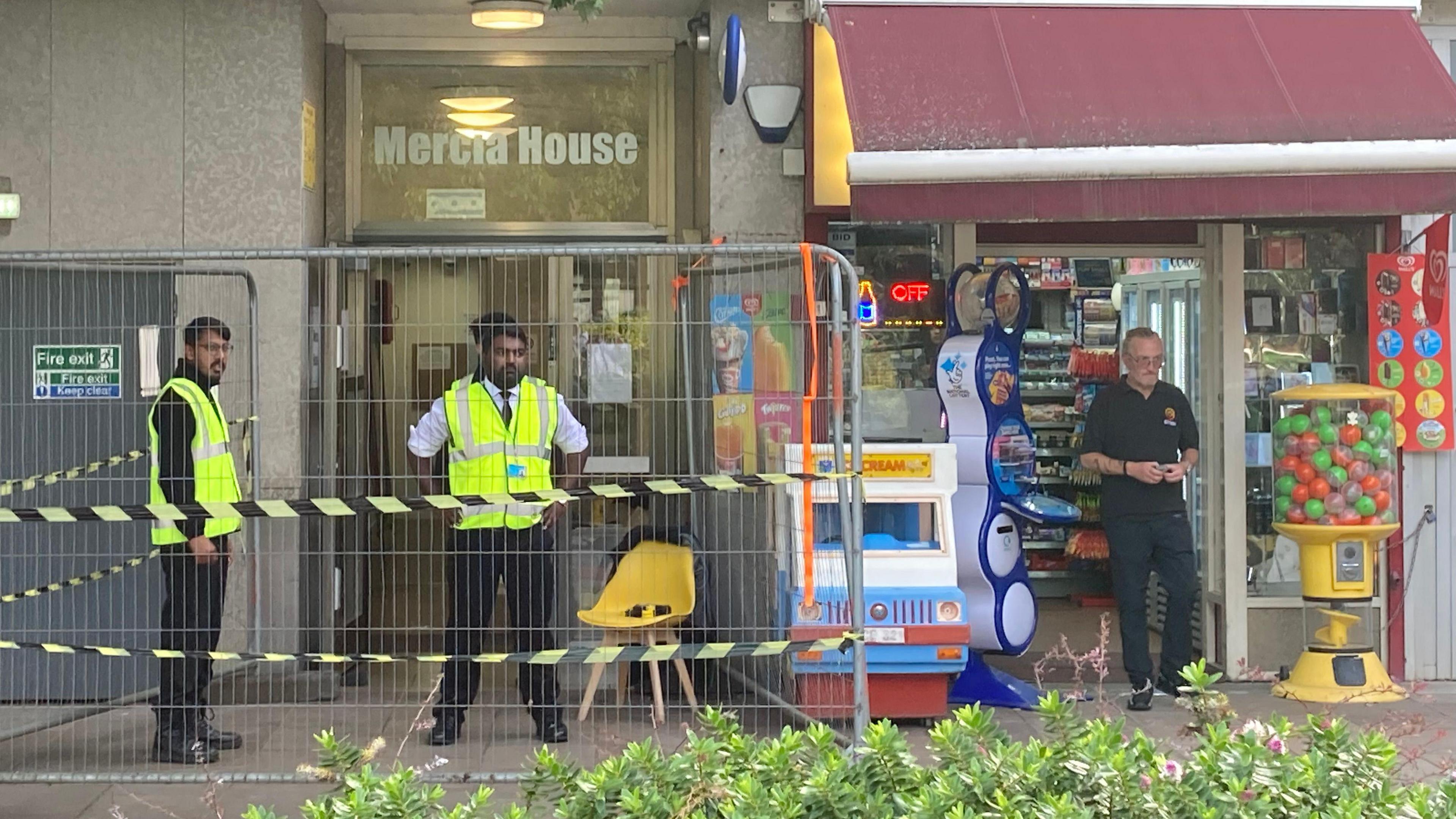 Security guards stand outside the entrance to Mercia House which is cordoned off with tape and barriers. There is a man standing in the doorway of the neighbouring shop.