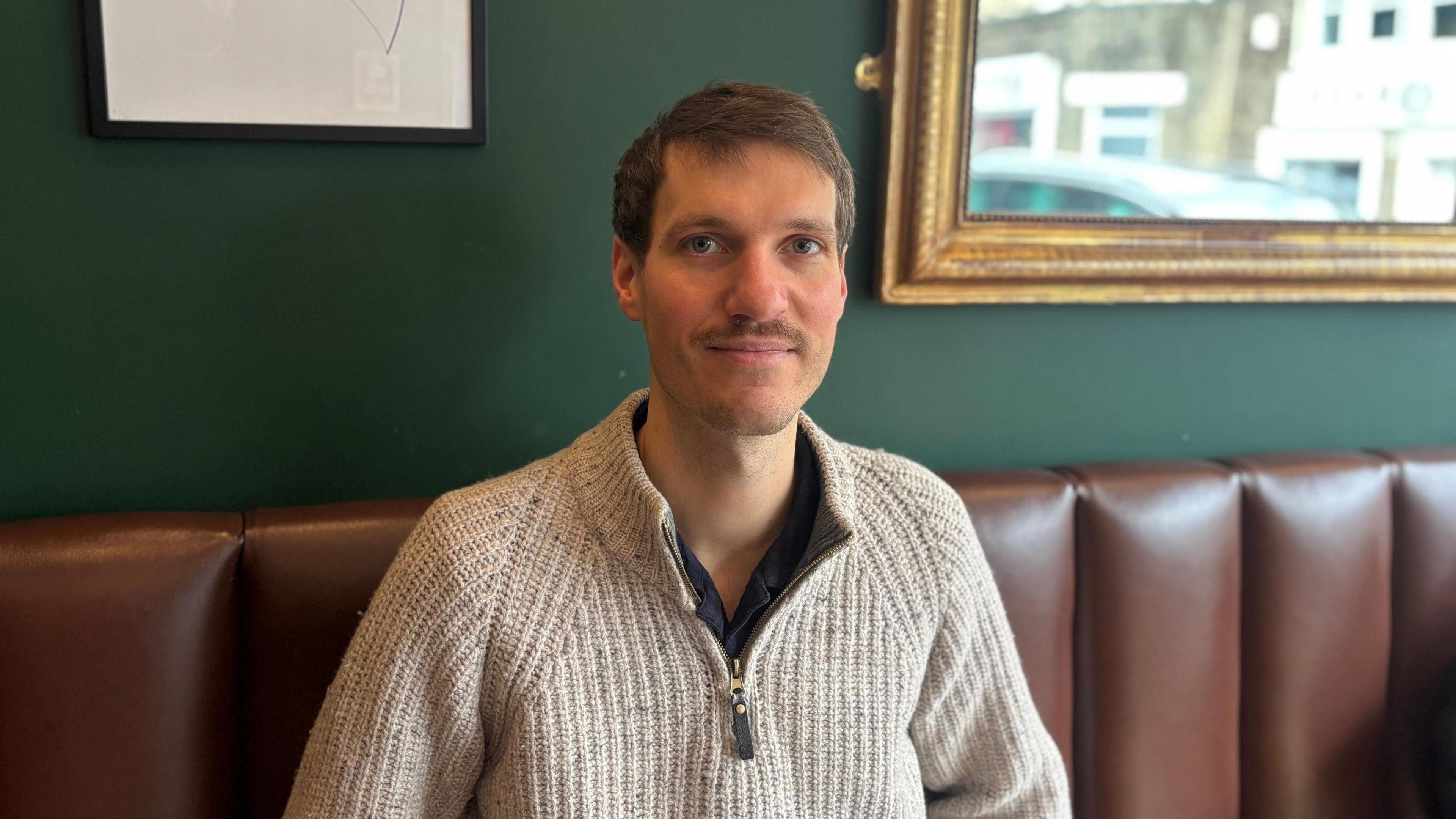 Clément Cousin is sat inside Bavette restaurant on a brown leather bench. He has a green wall behind him with a mirror on it. He has brown hair and a moustache, and is wearing a cream quarter-zip knitted fleece.