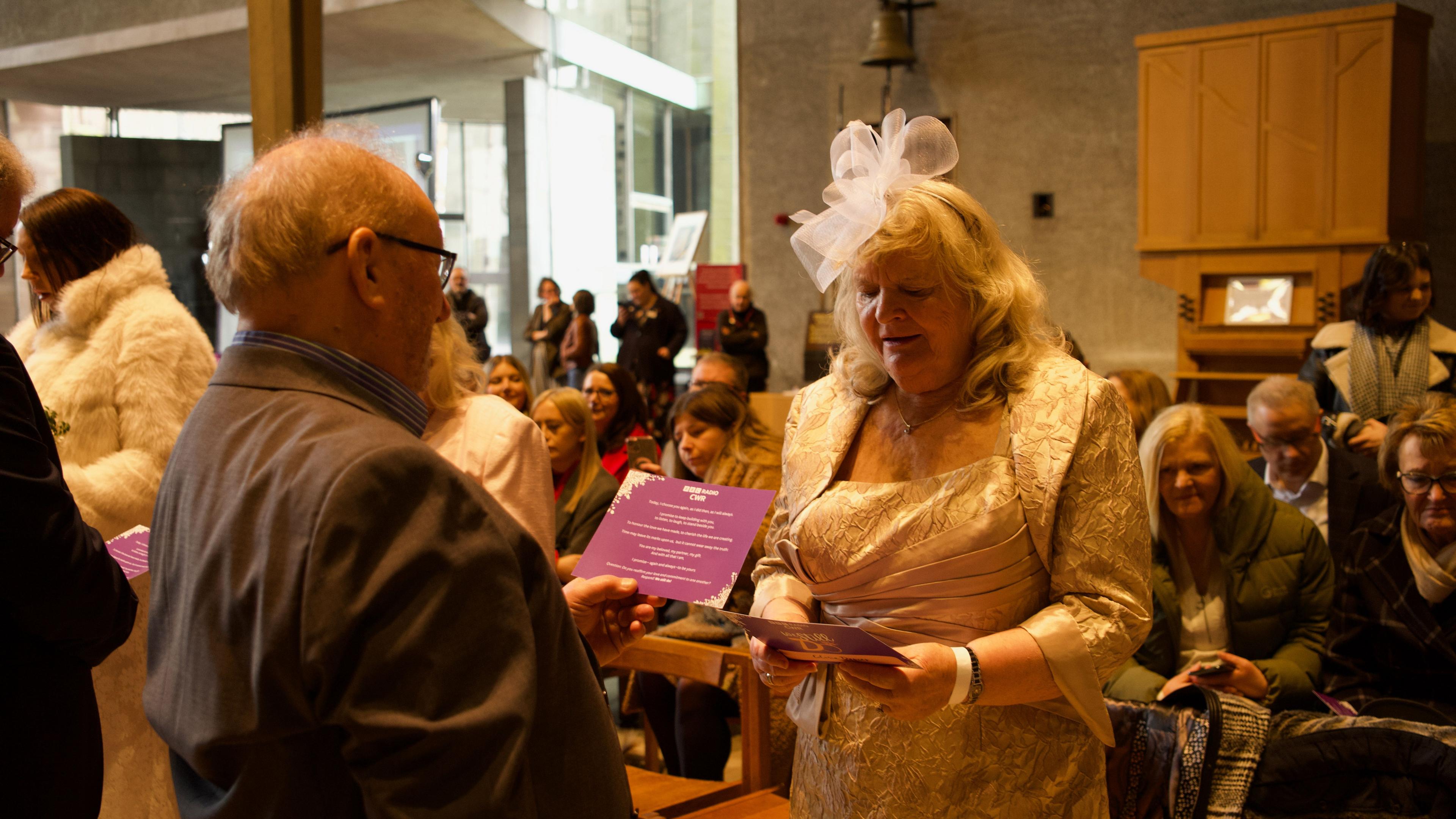 A man in a suit and woman in a golden dress and jacket with white fascinator read from purple cards during the event