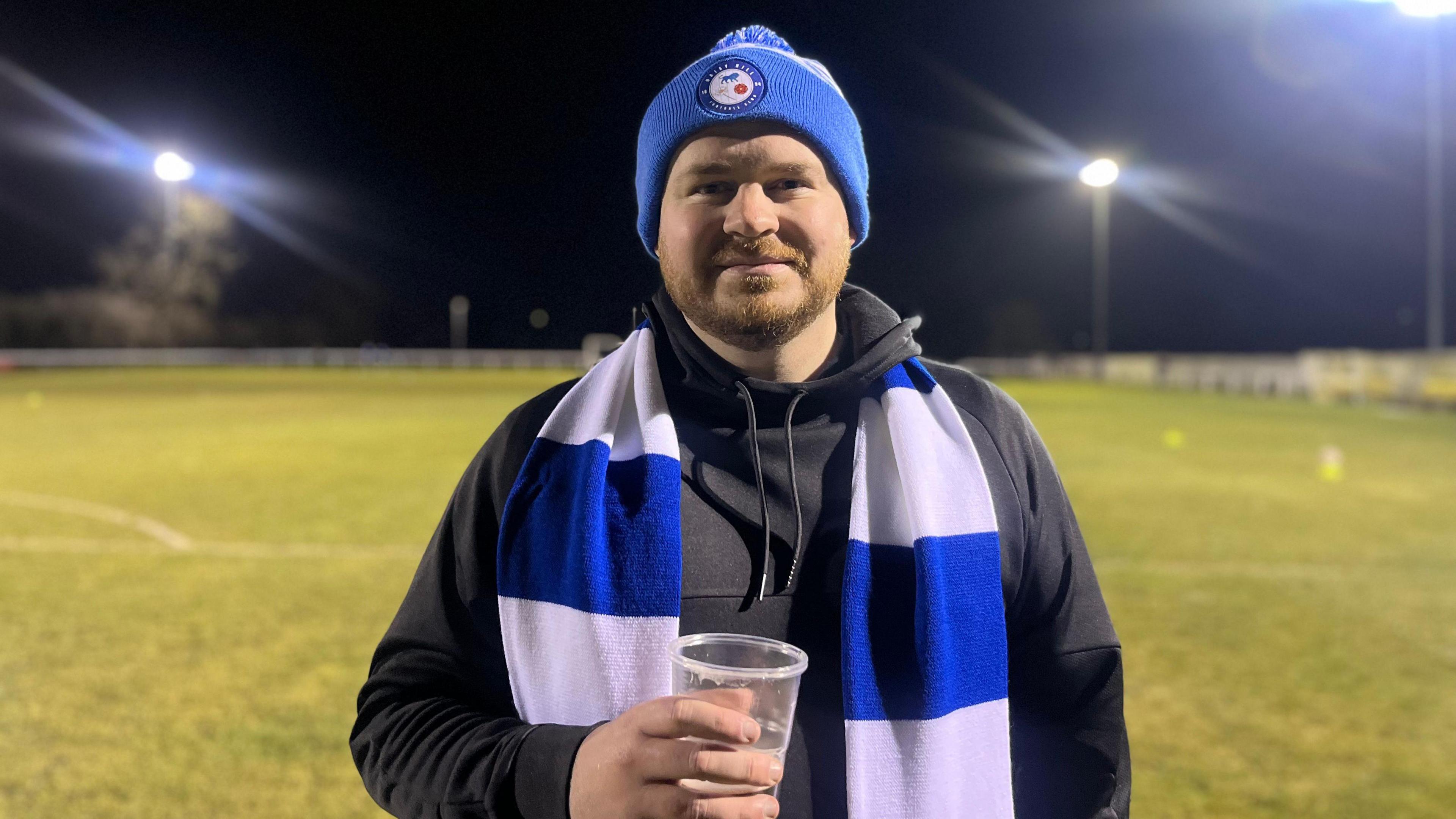 Aaron Hunt, wearing a blue and white woolly hat and scarf over a black hoodie, smiles at the camera holding a pint of beer in a plastic cup. He is standing on a football pitch illuminated with floodlights. 