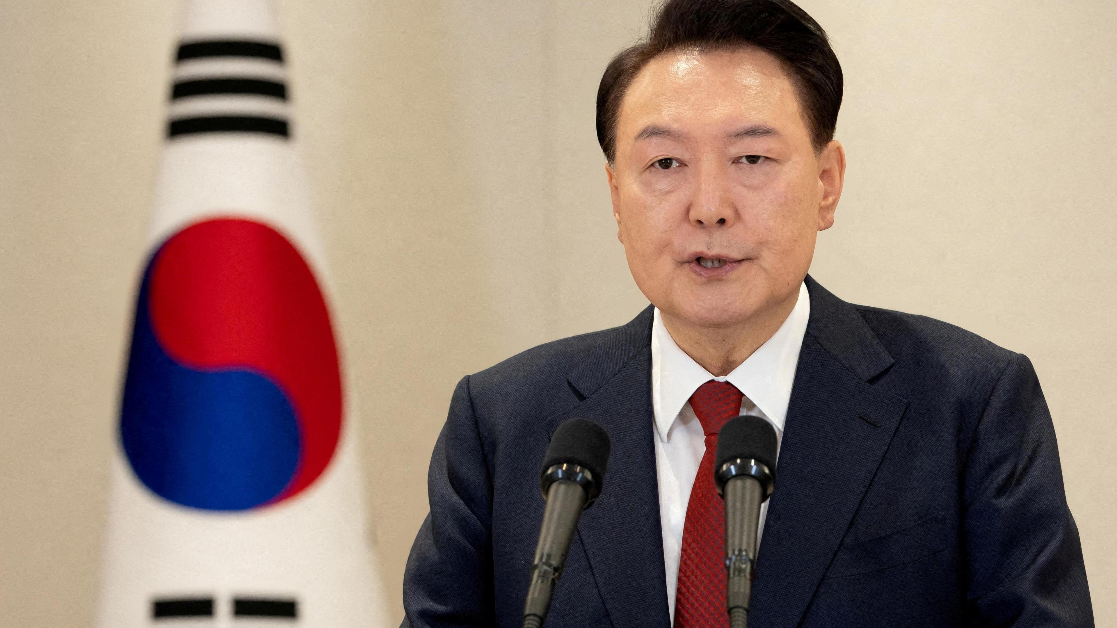 South Korean President Yoon Suk Yeol delivers an address to the nation at the Presidential Office in Seoul, South Korea. He wears a dark blazer, a red tie and stands to the side of the South Korean falg. 